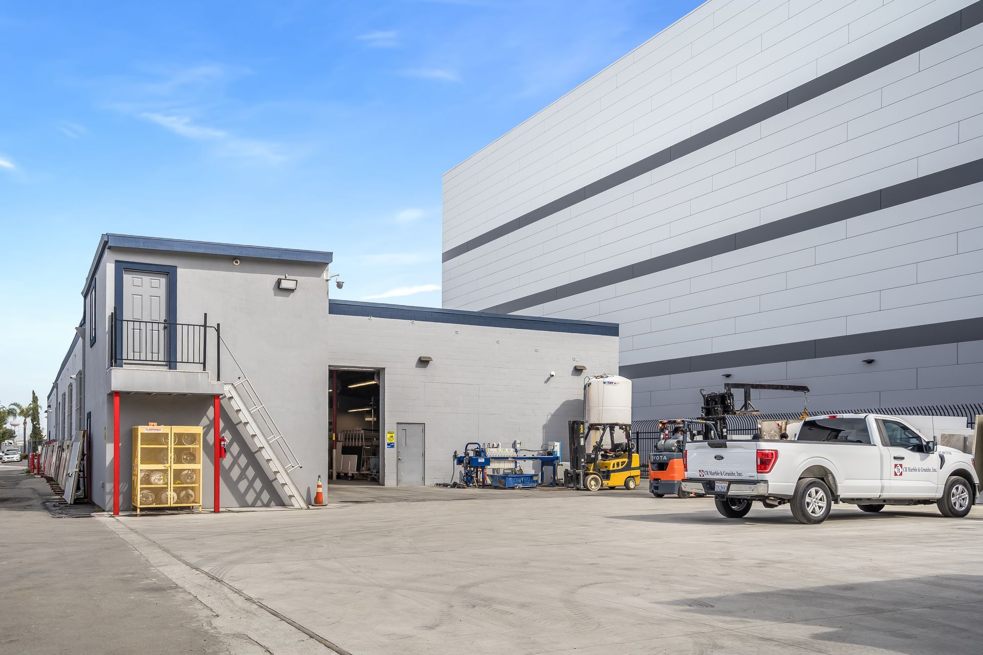 Industrial yard with a small building, parked trucks, and warehouse-style structures under a blue sky
