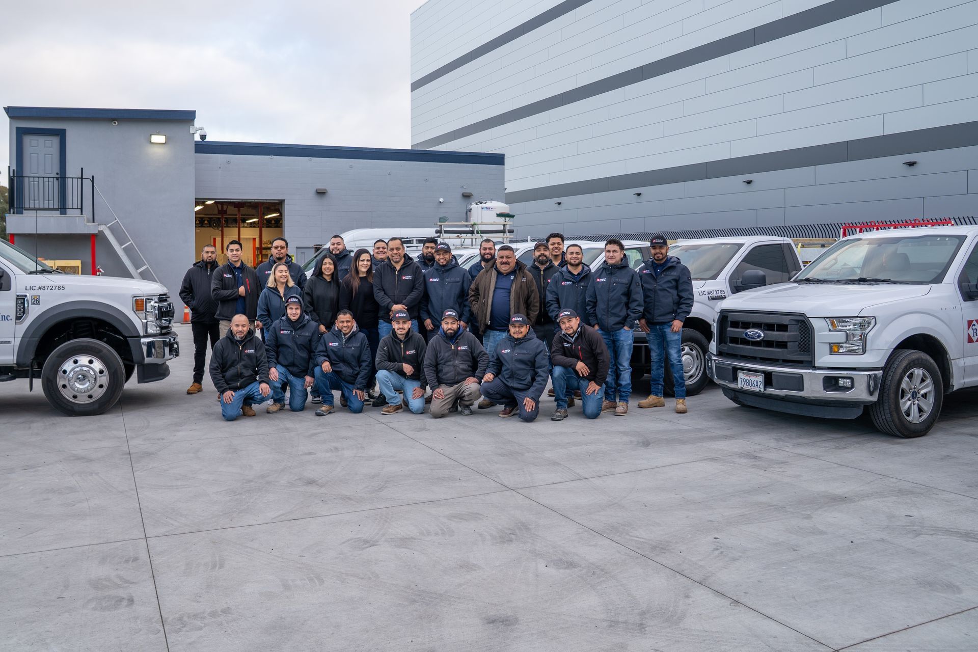 Group of people posing outdoors between two white pickup trucks beside a large industrial building