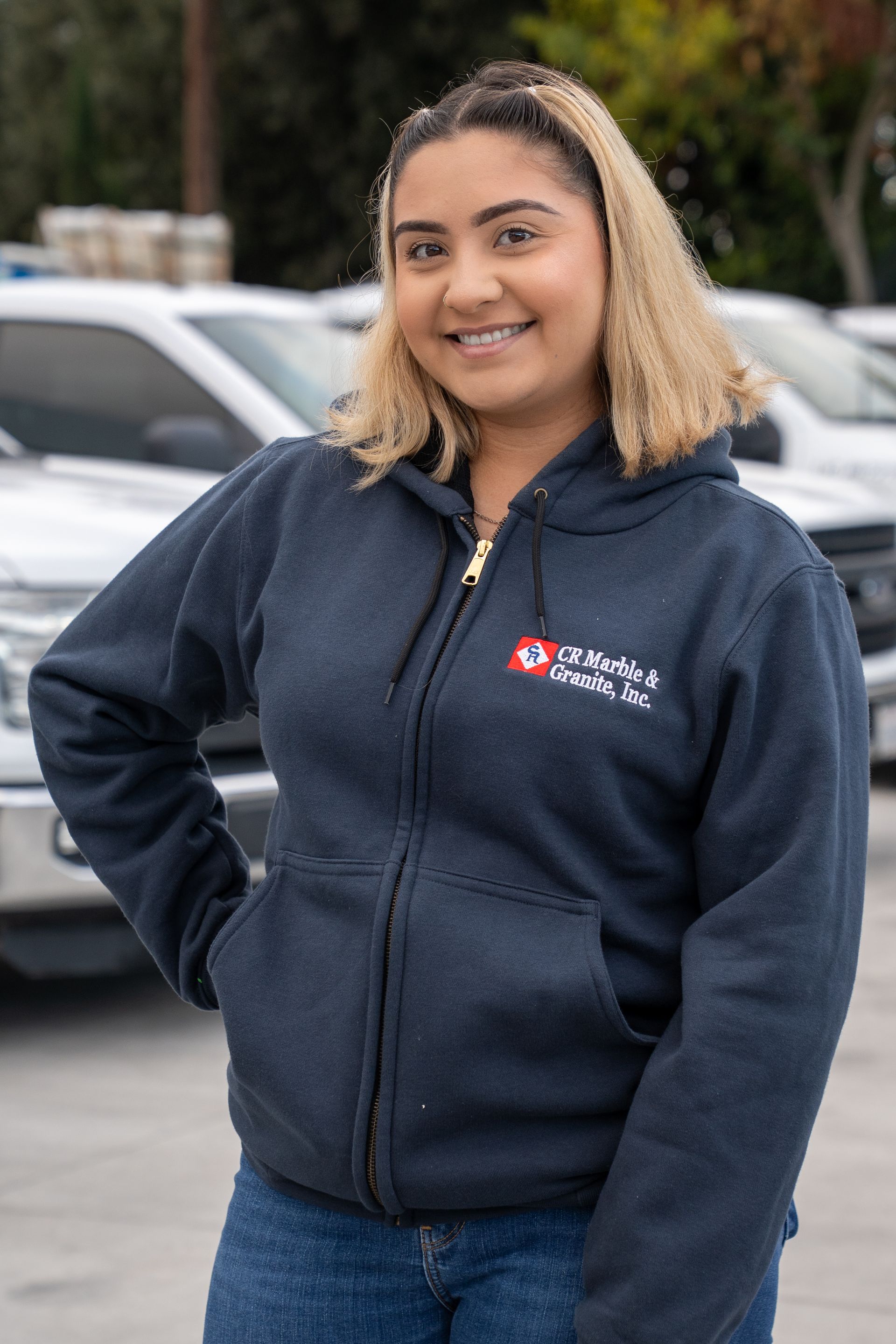 Smiling person in a navy zip-up hoodie standing outdoors in a parking lot.