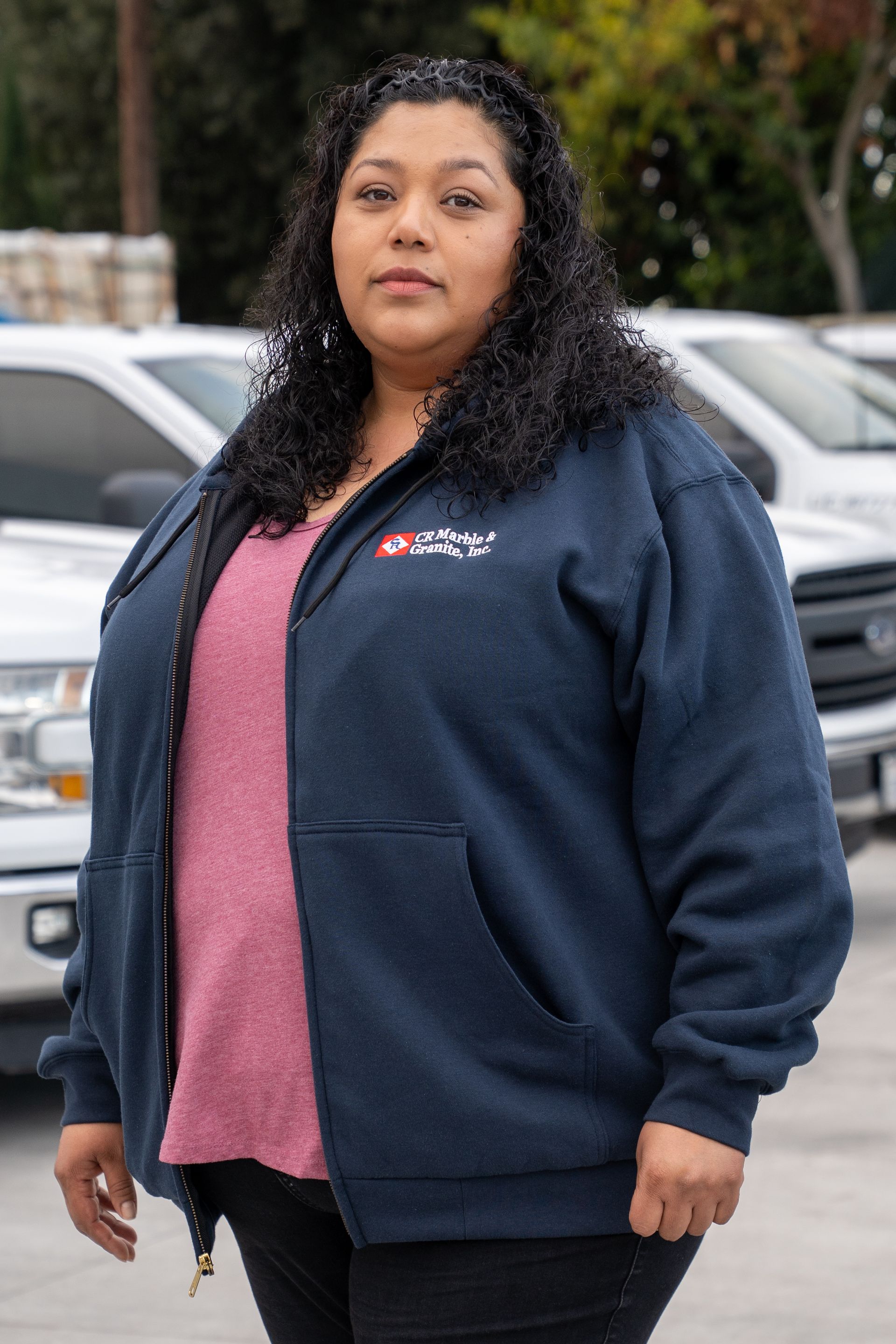 Woman standing outdoors in a navy jacket and pink shirt, with parked cars in the background
