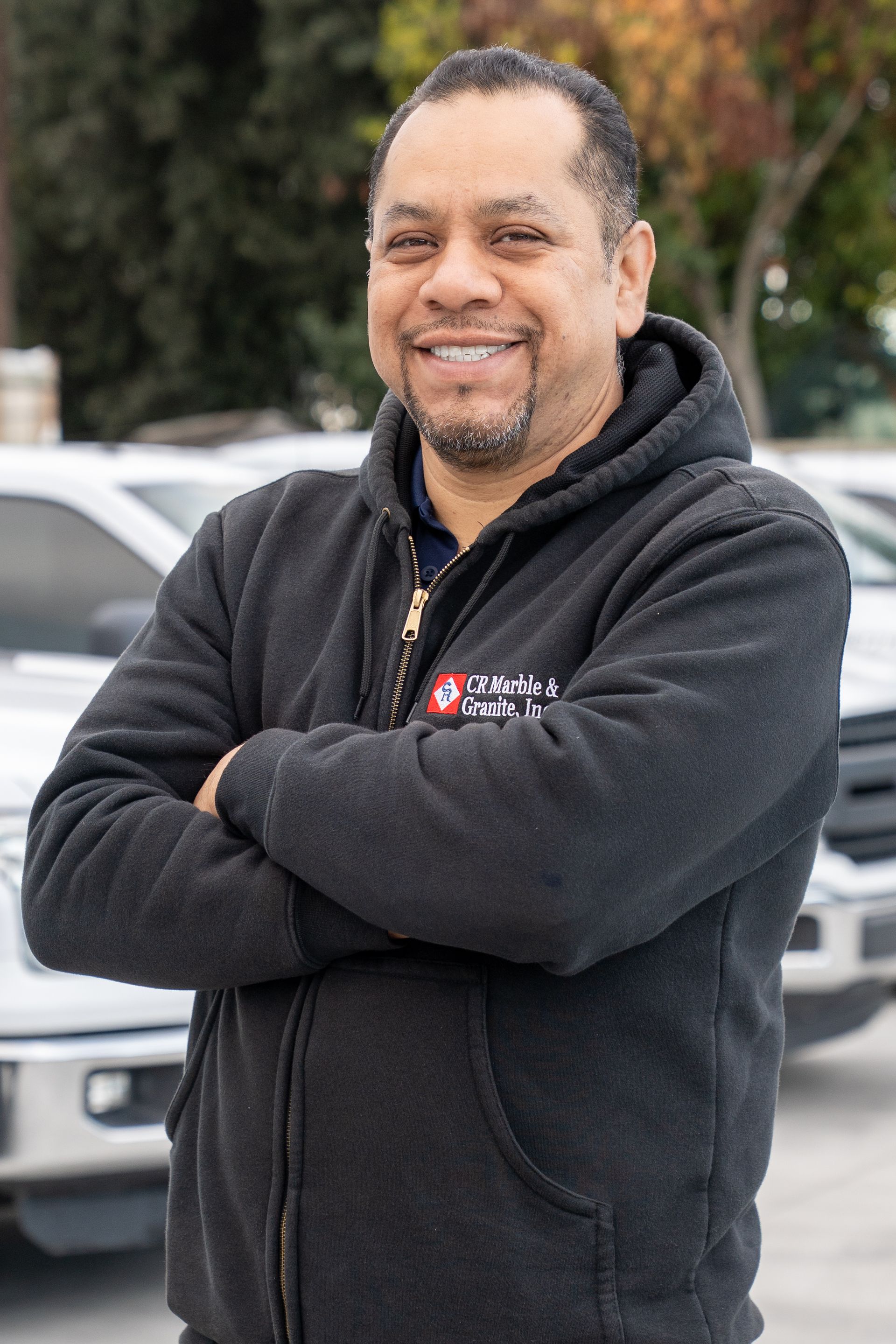 Smiling man in a black hoodie with arms crossed, standing outdoors in front of a vehicle.