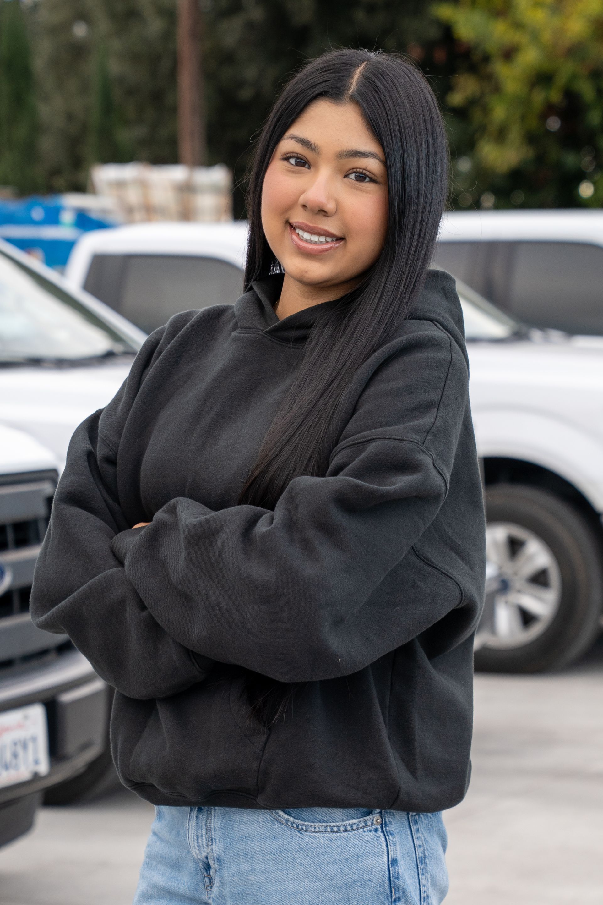 Person in a black hoodie stands with arms crossed in front of parked cars outdoors.