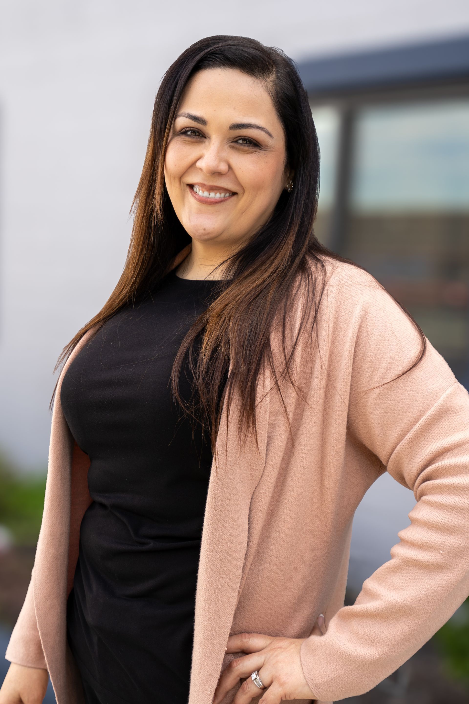 Smiling woman in a black top and pink cardigan standing outdoors near a building