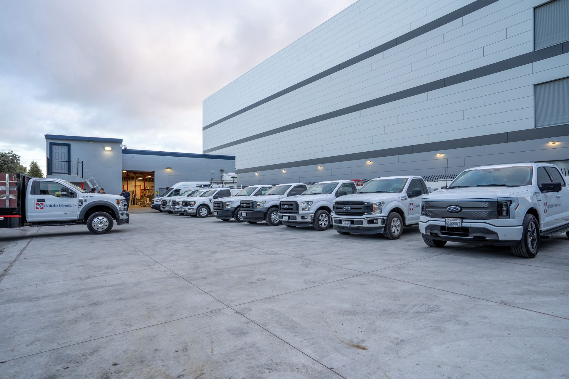 Line of white trucks parked outside a large industrial building at dusk.