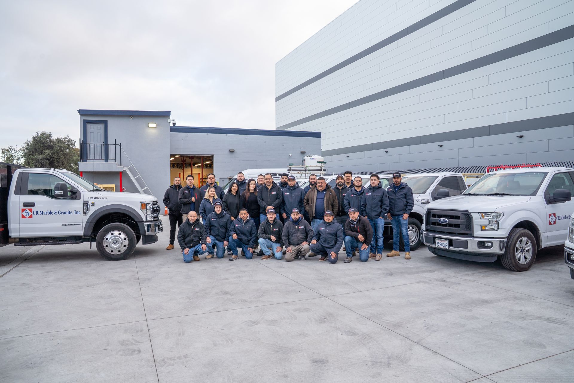 Group of uniformed workers posing with two white trucks outside a warehouse.