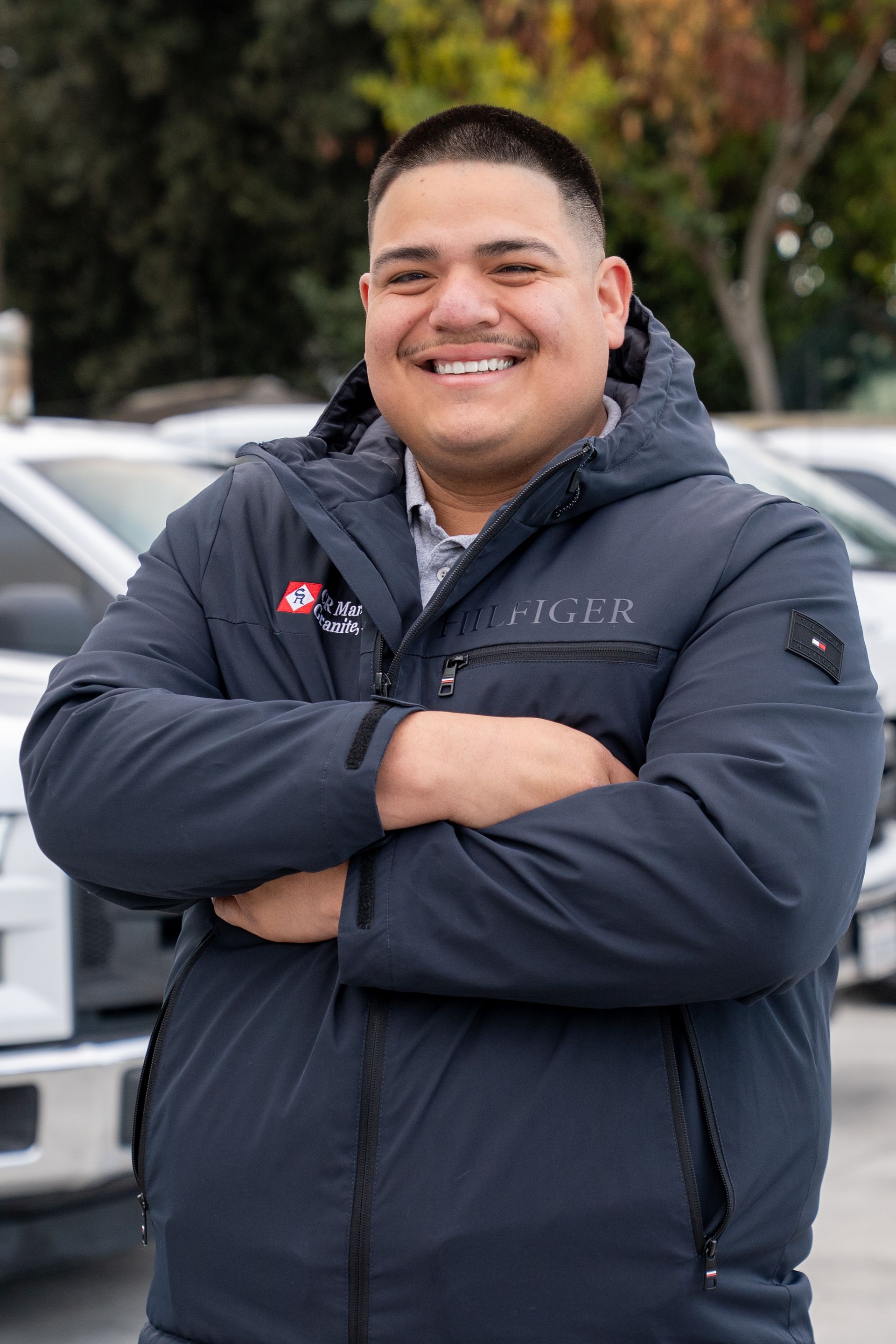Smiling person in a navy puffer jacket with arms crossed, standing outdoors in front of cars.