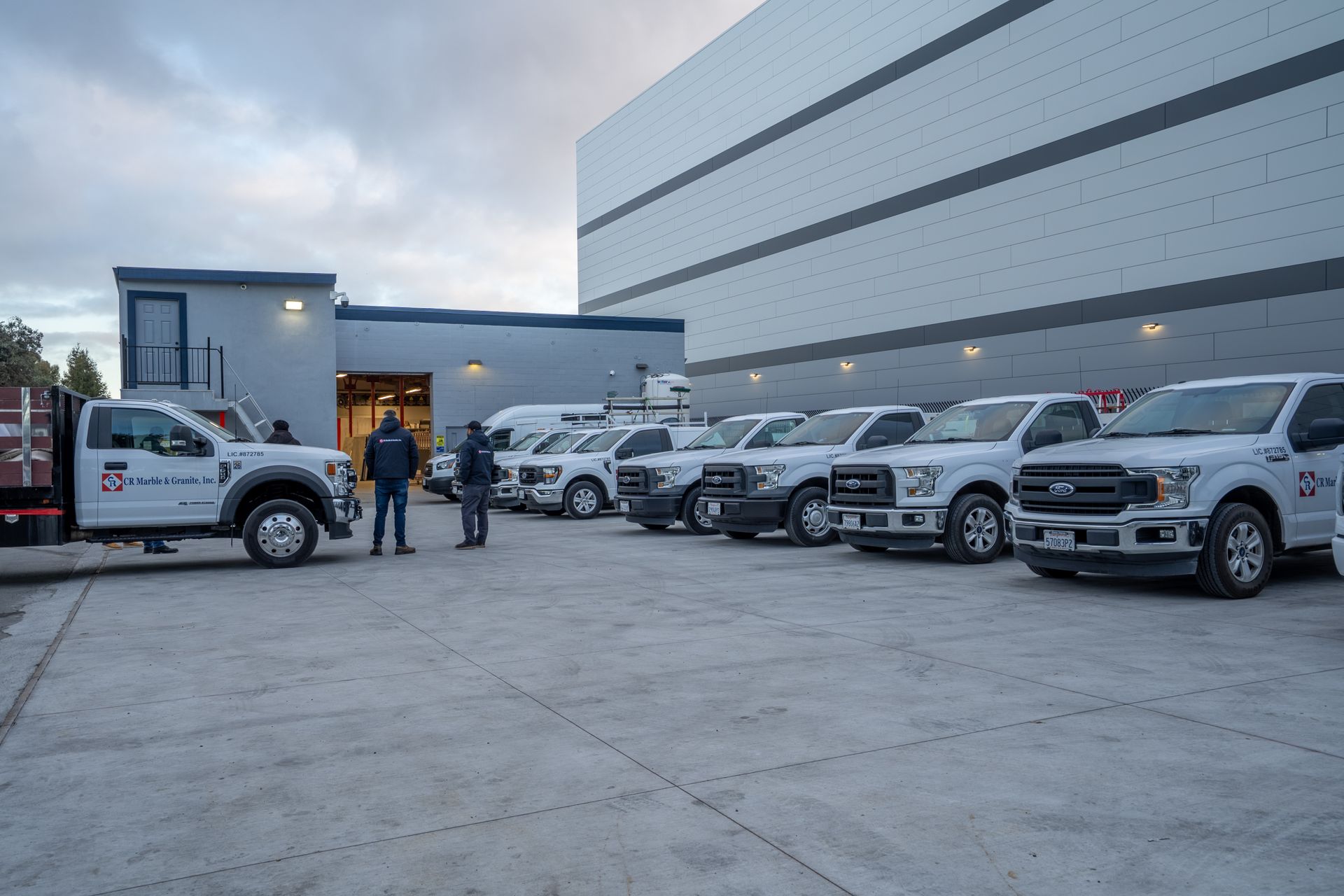 Fleet of white work trucks parked outside a warehouse at dusk, with two people standing near the entrance