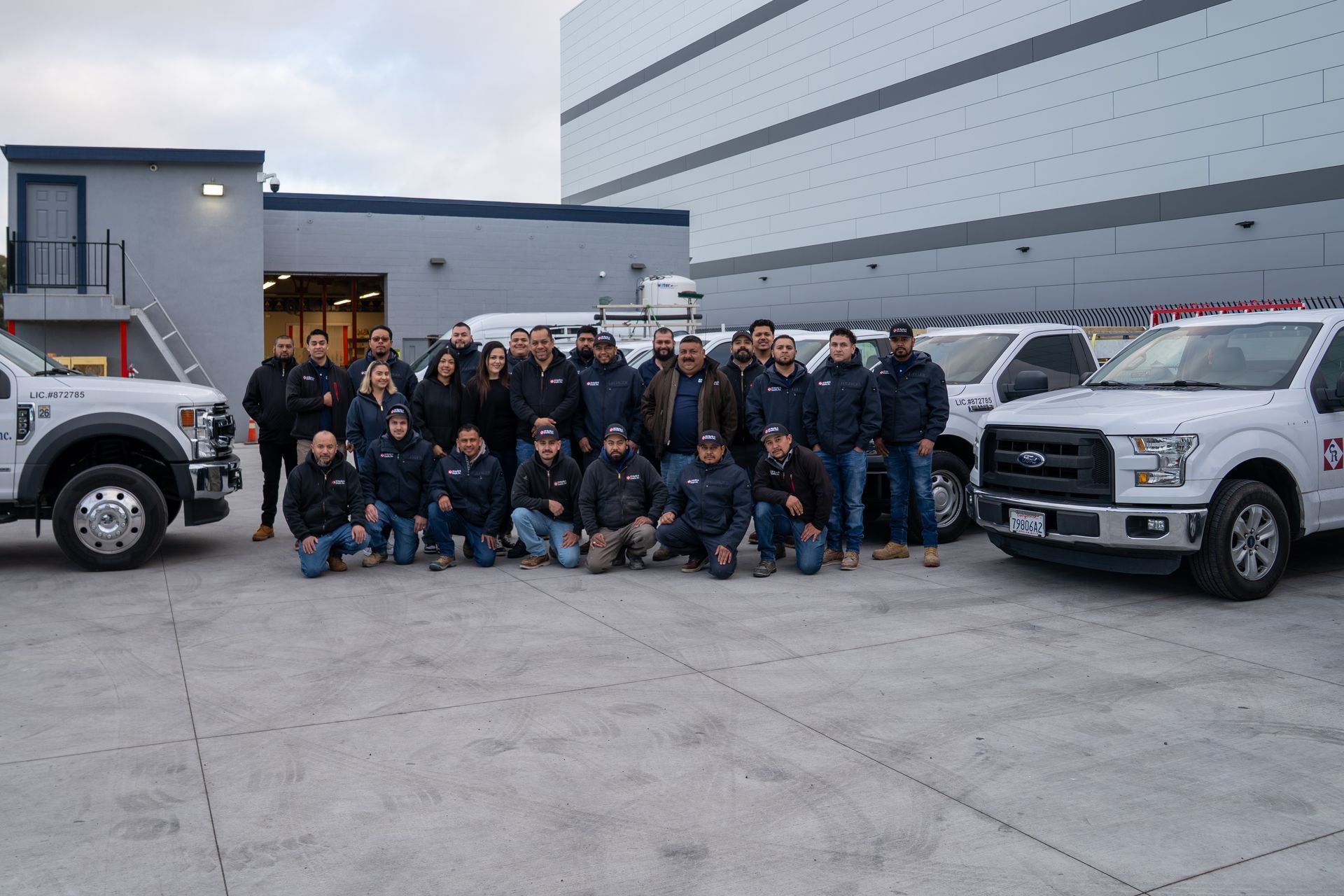 Group of uniformed workers posing between two white trucks in an industrial parking lot