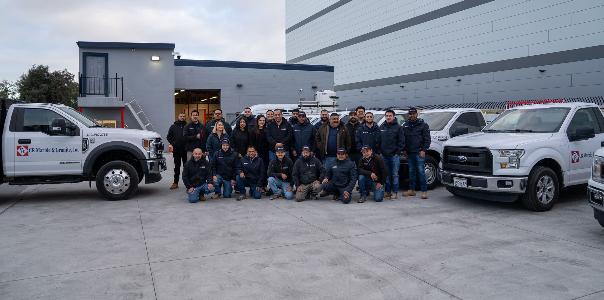Group of utility workers posing between two white pickup trucks outside a building at dusk
