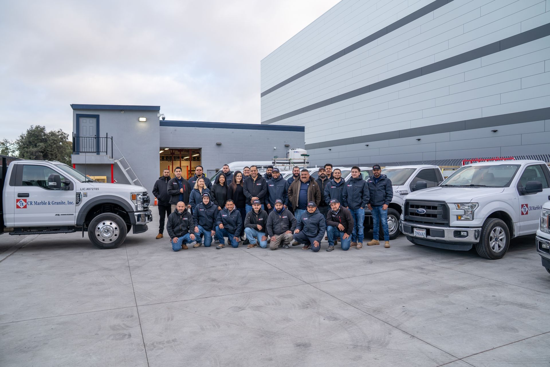 Group of workers posing outside an industrial building beside two white service trucks