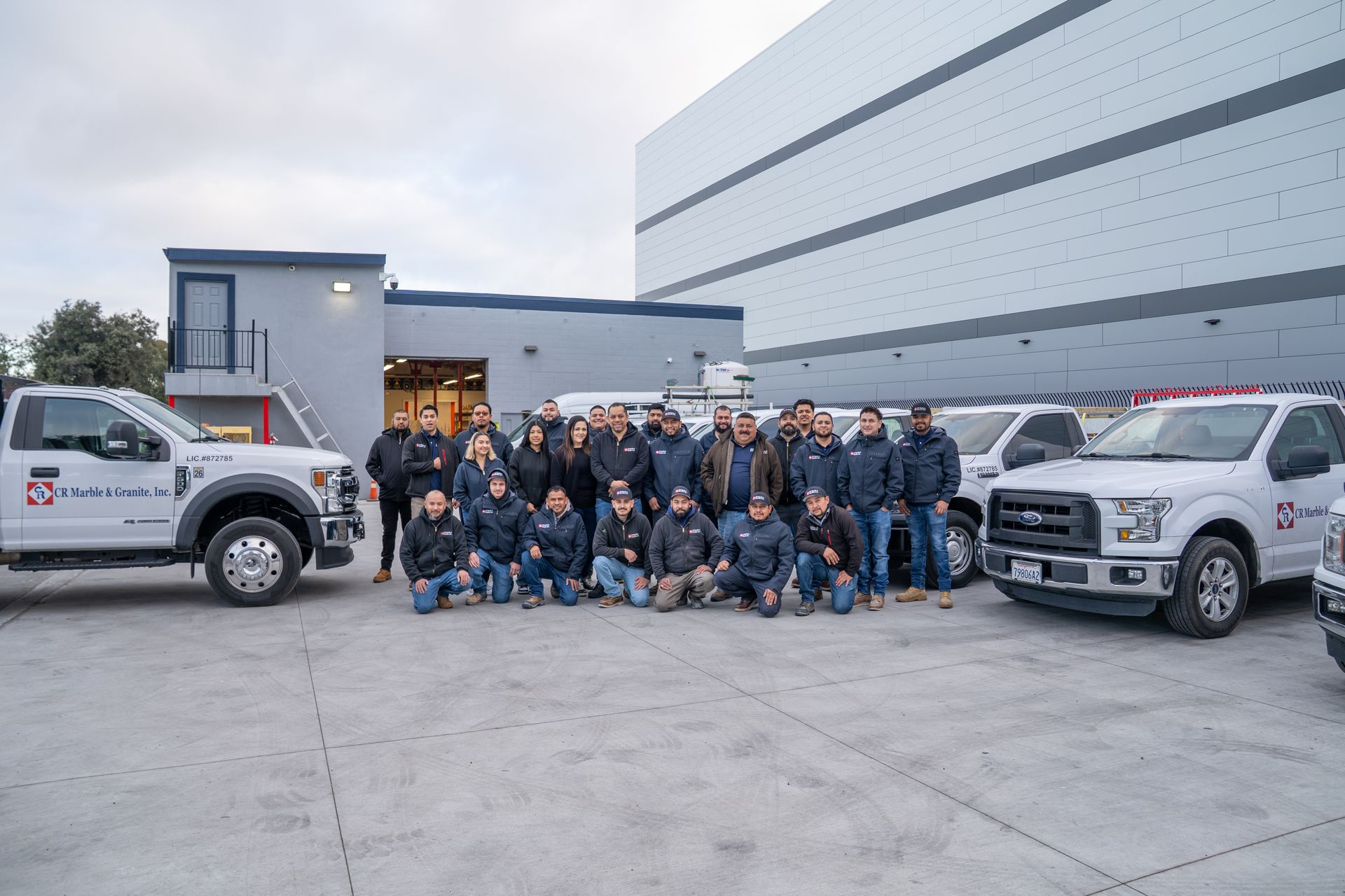 Group of workers posing in front of white trucks outside a warehouse on a concrete lot