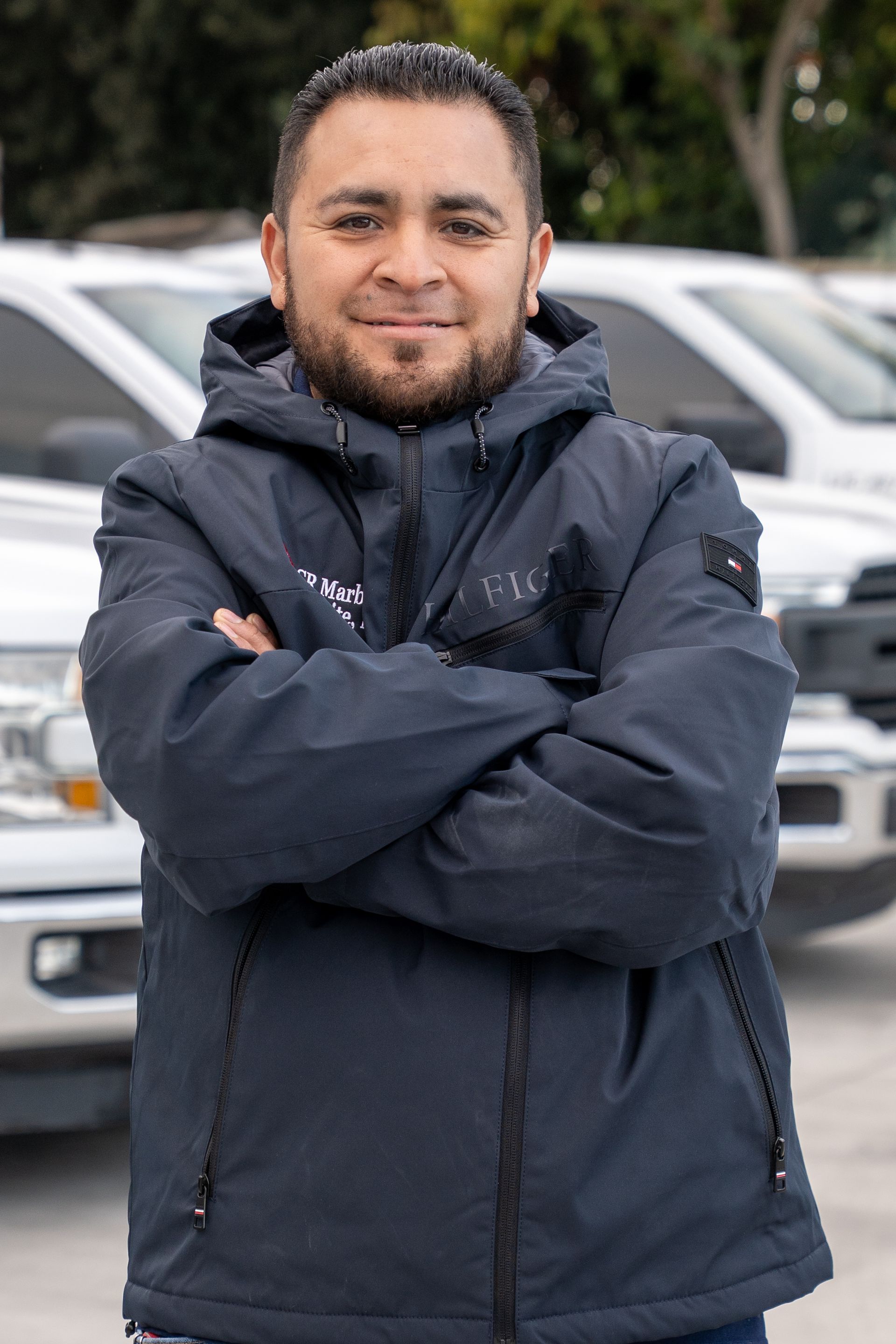 Person standing with arms crossed in a dark jacket in front of parked cars.