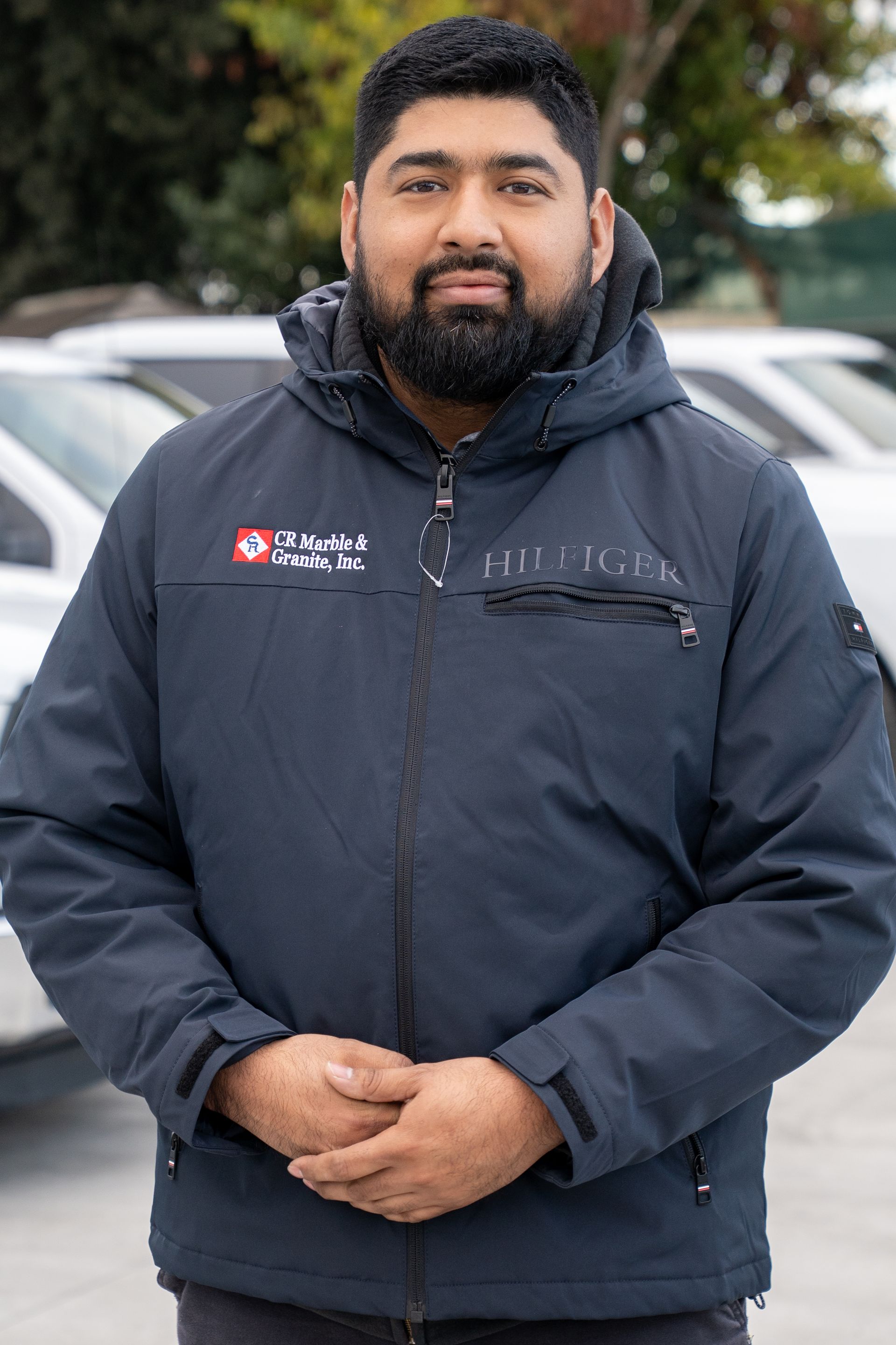 Man in a dark jacket standing outdoors in front of parked cars, hands clasped, with a neutral expression