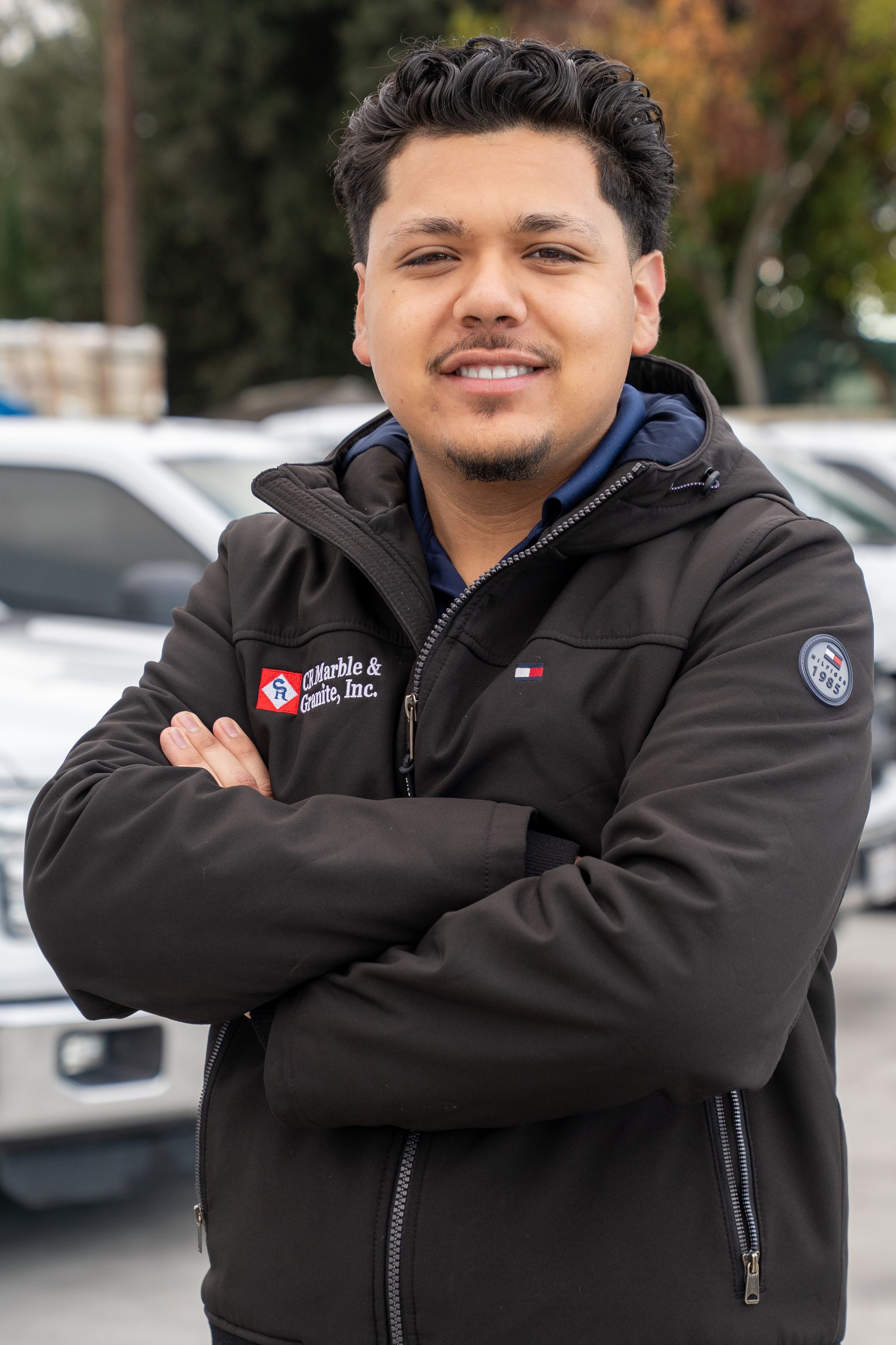 Man standing with arms crossed in a black jacket outdoors, with cars and trees in the background