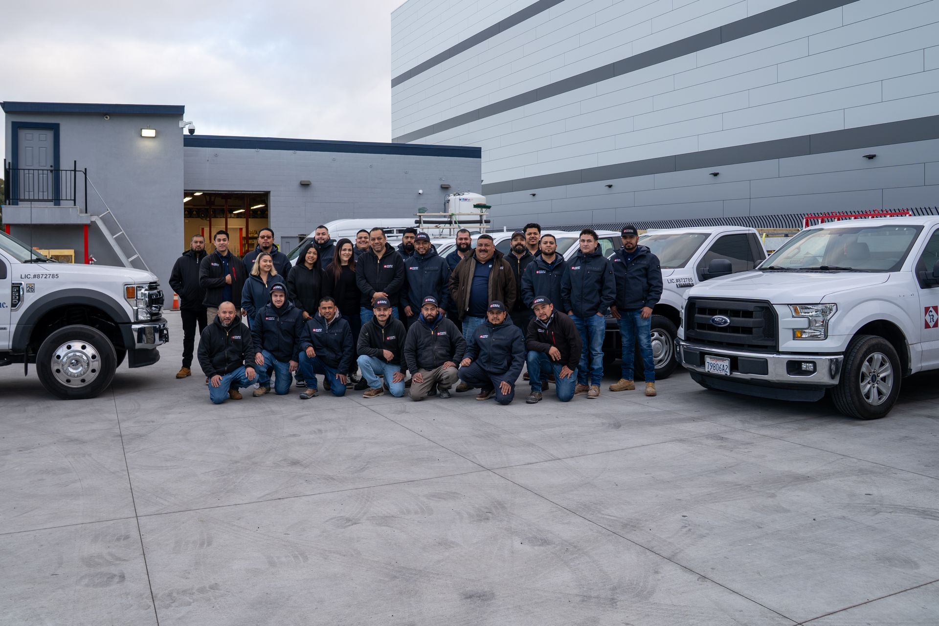Group of people posing outdoors between two white pickup trucks in front of a large building.