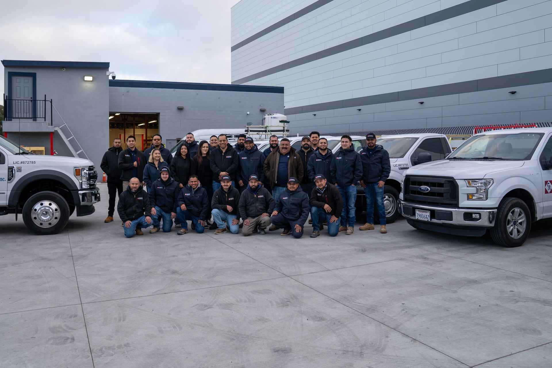 Group of people in dark uniforms posing between two white pickup trucks outside a building.