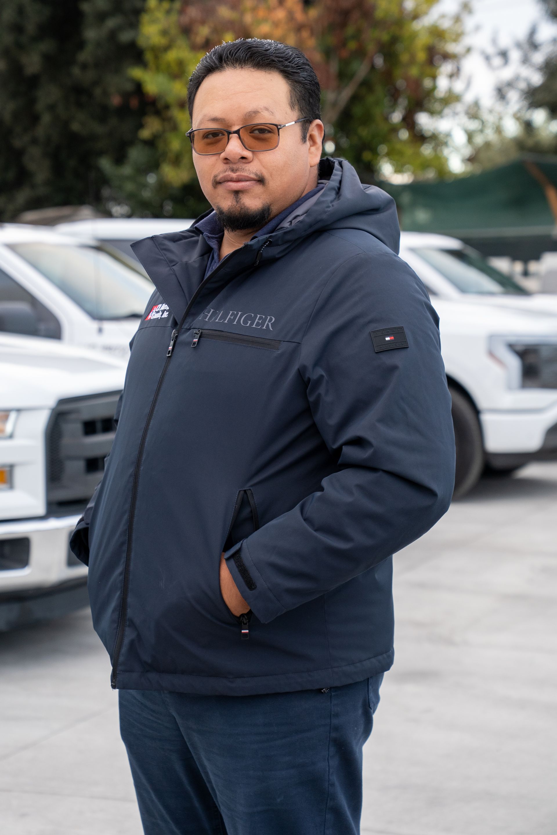 Man in a navy jacket and jeans standing outdoors near parked white vehicles.