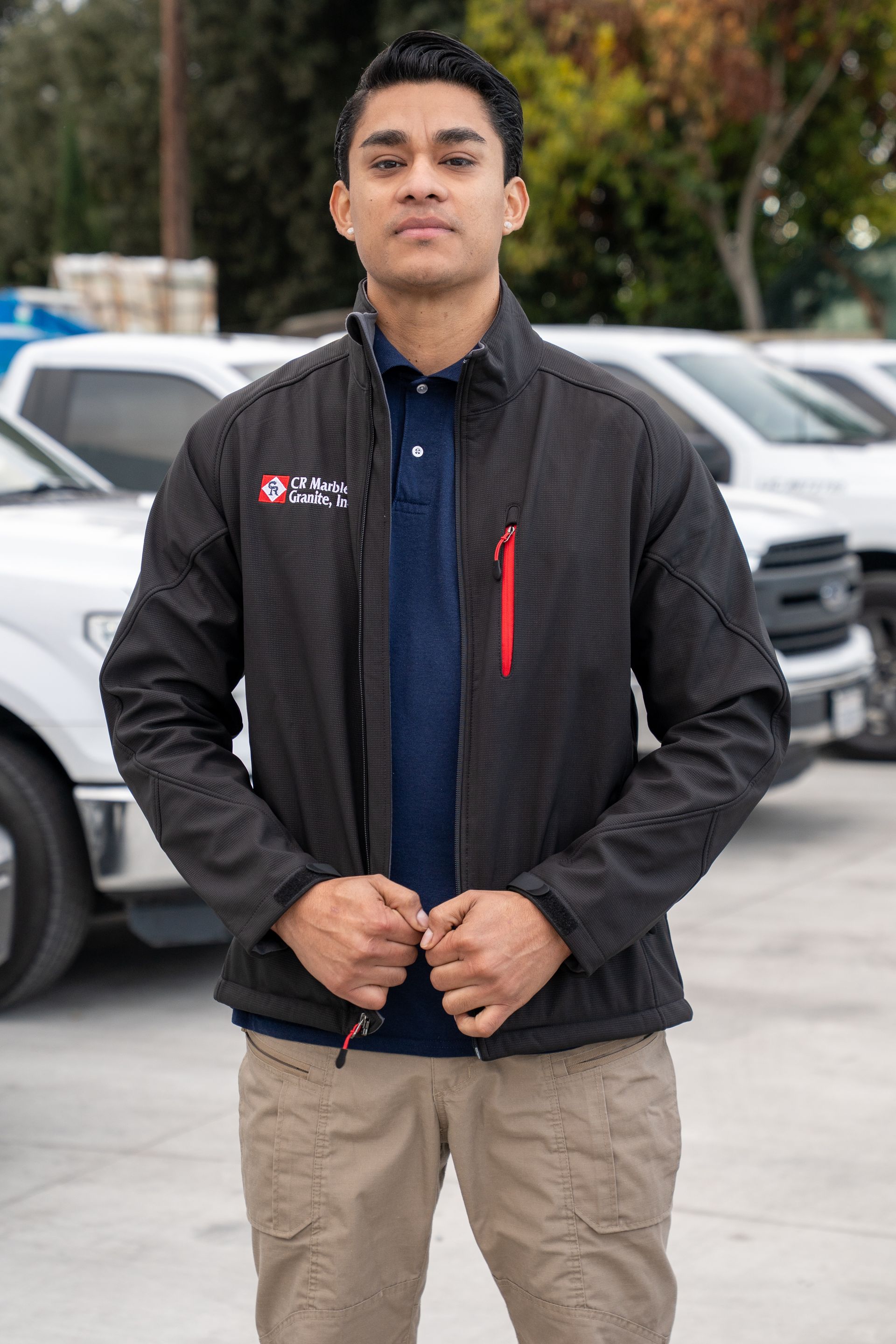Person standing outdoors in a black jacket and tan pants, holding the jacket zipper closed.