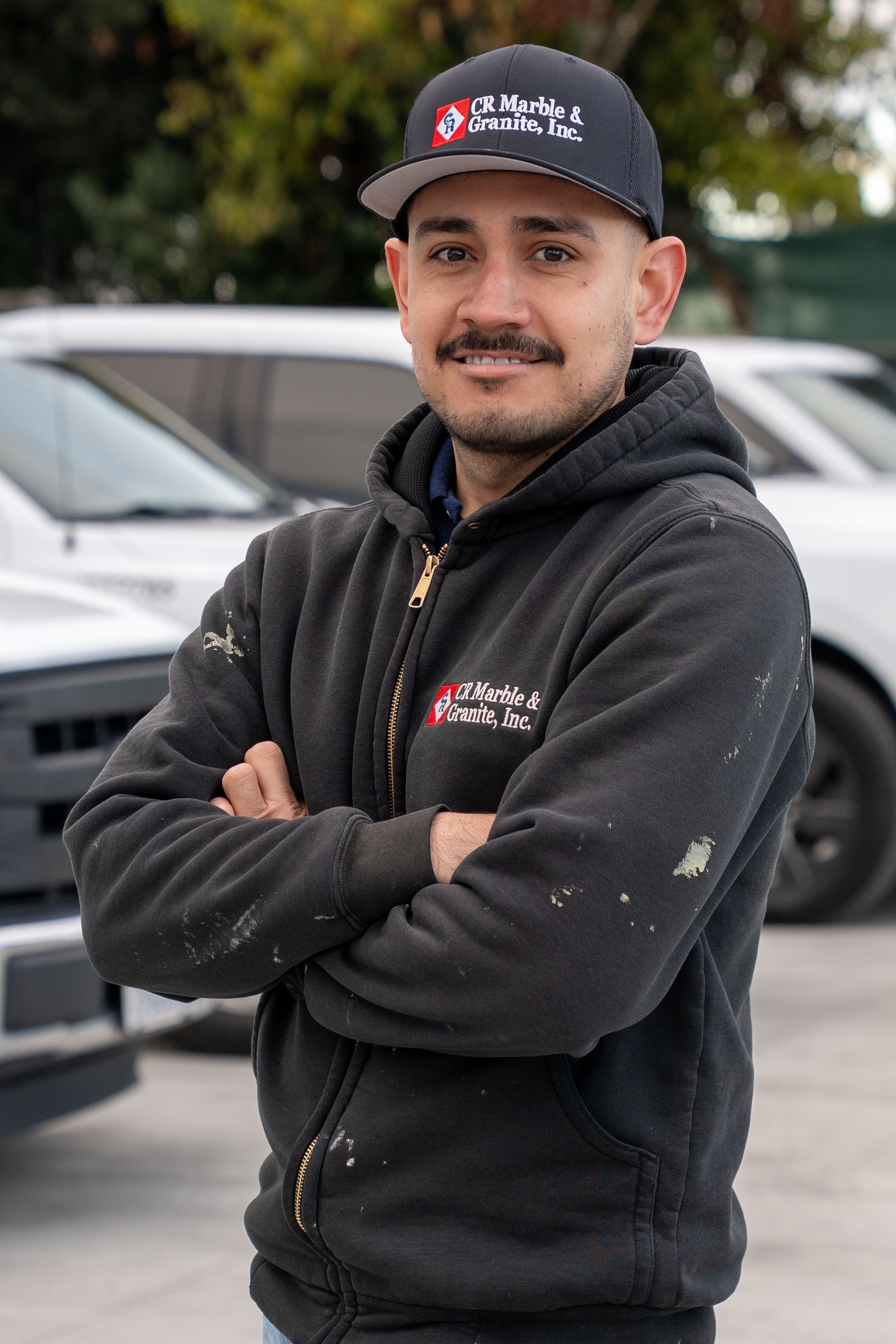 Man standing with arms crossed in a black cap and hoodie in front of parked cars