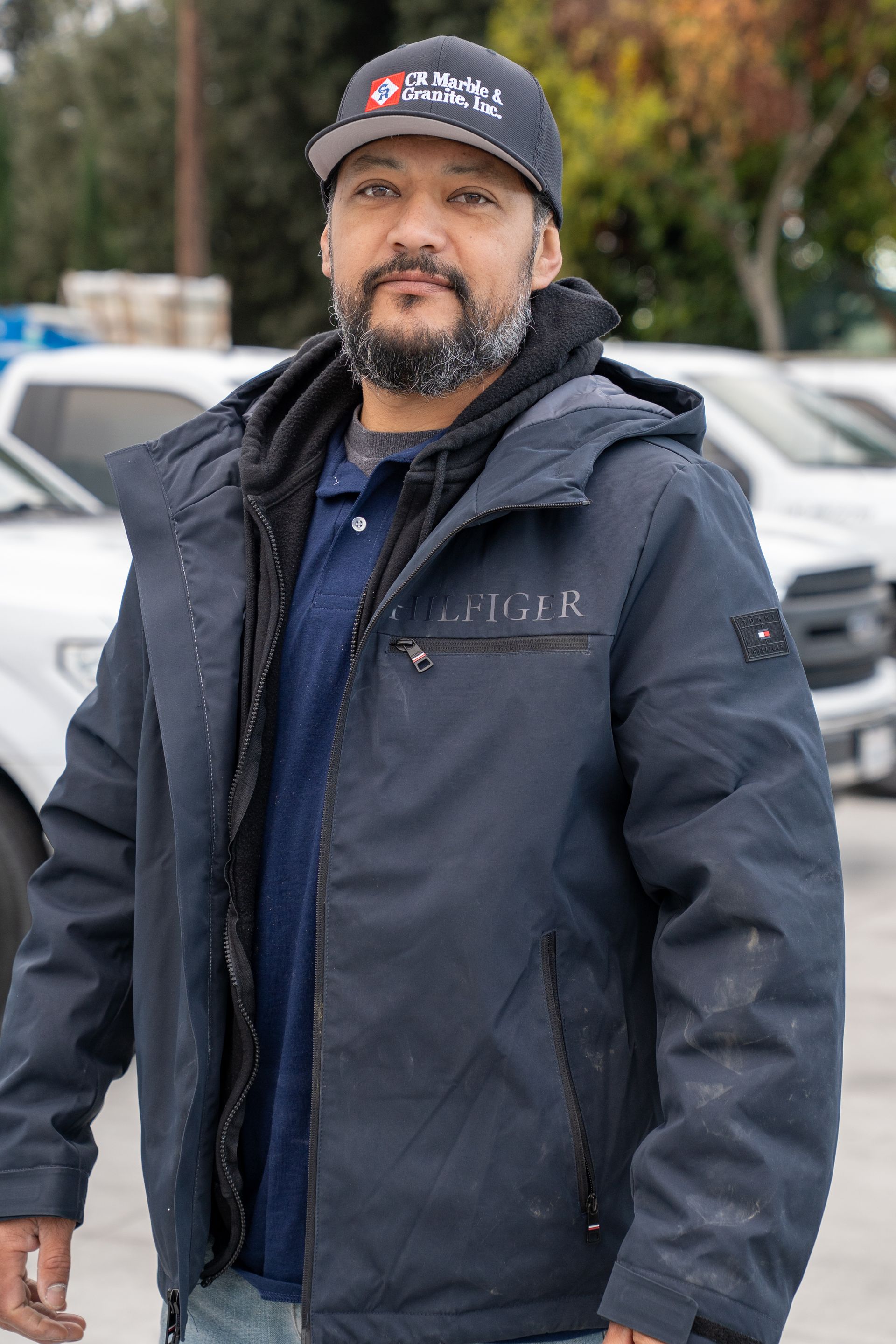 Man in a navy jacket and cap standing outdoors with parked cars in the background