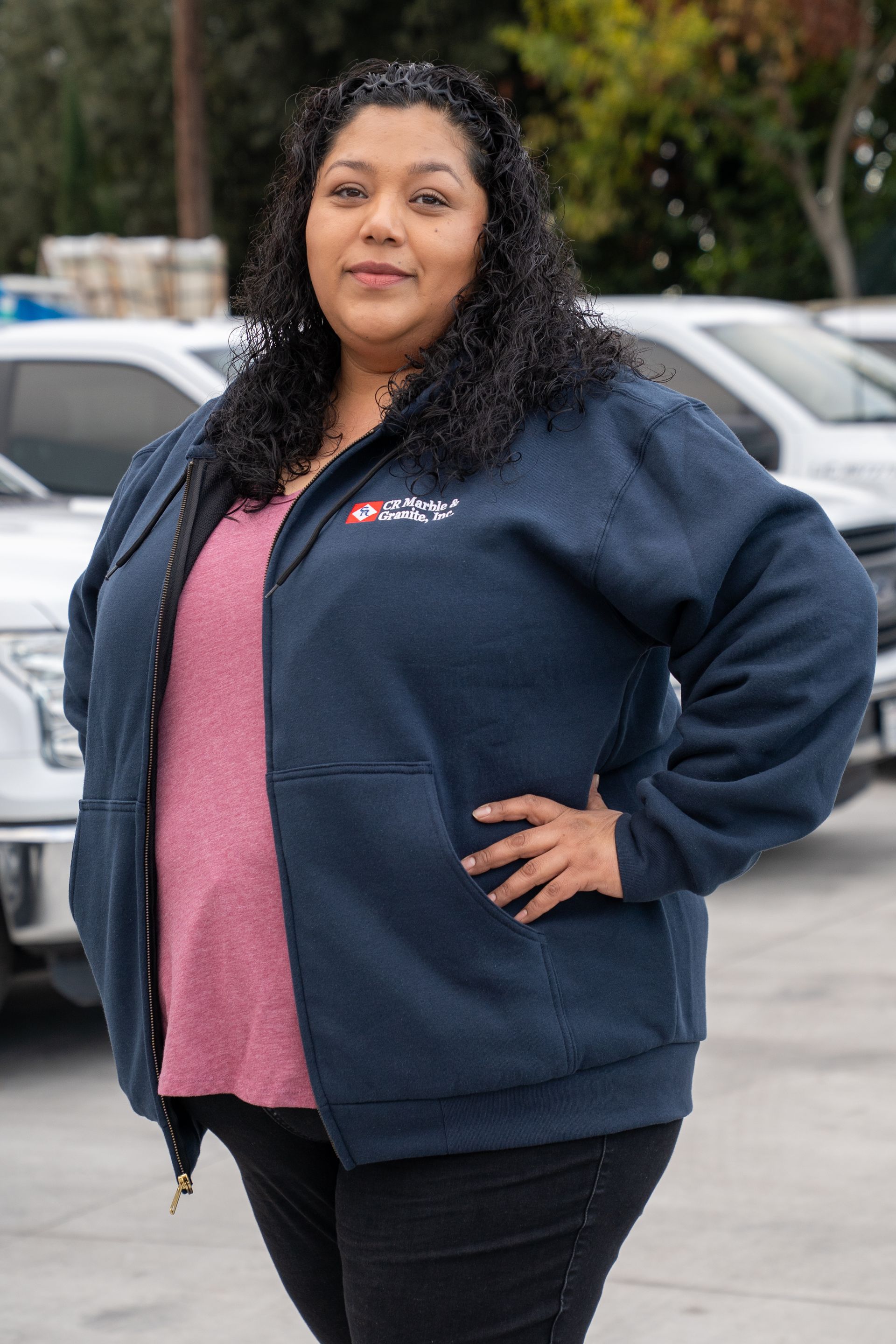 Woman standing outdoors with hands on hips, wearing a navy jacket over a pink shirt.