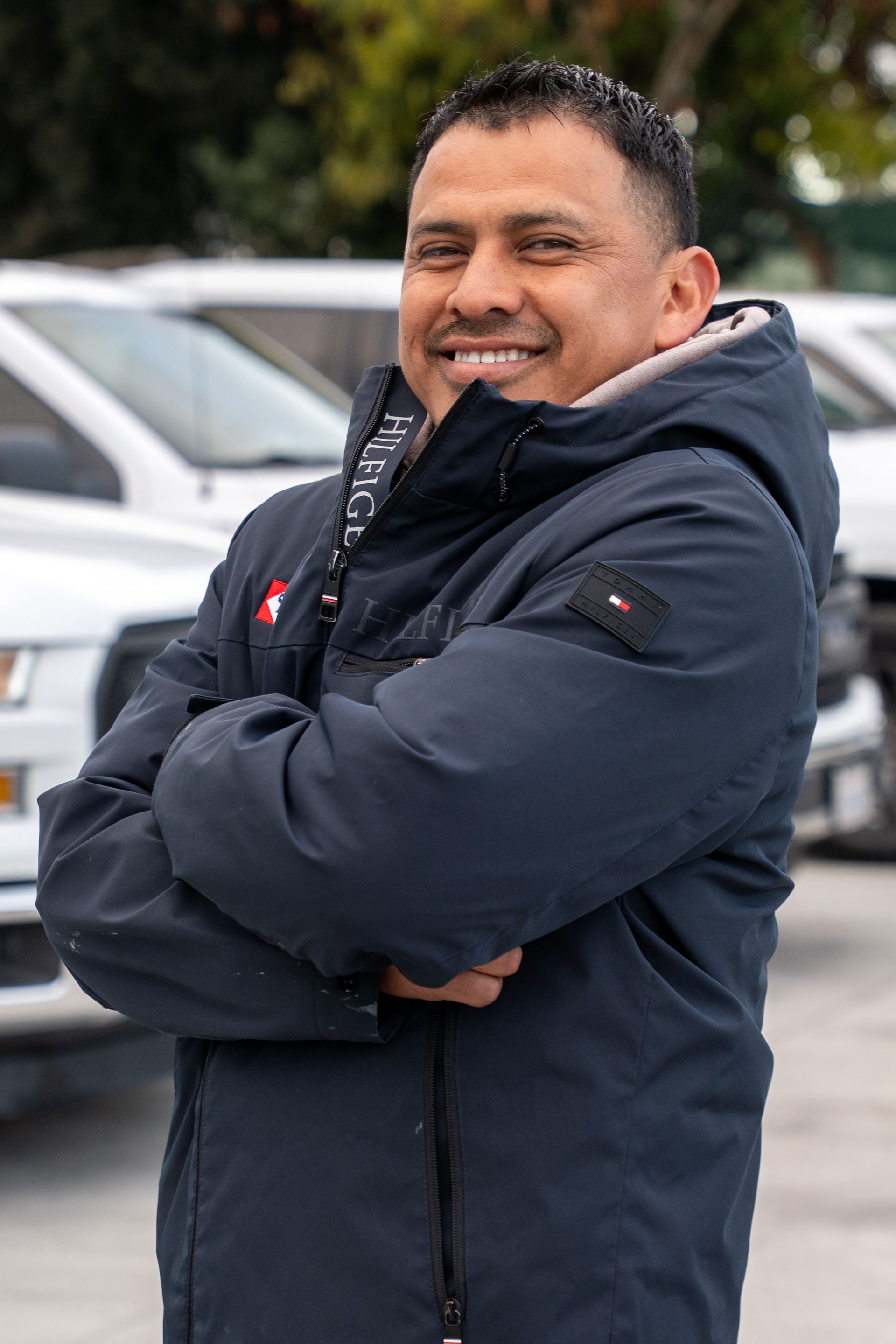 Smiling person in a dark jacket standing outdoors with arms crossed, parked cars blurred behind.