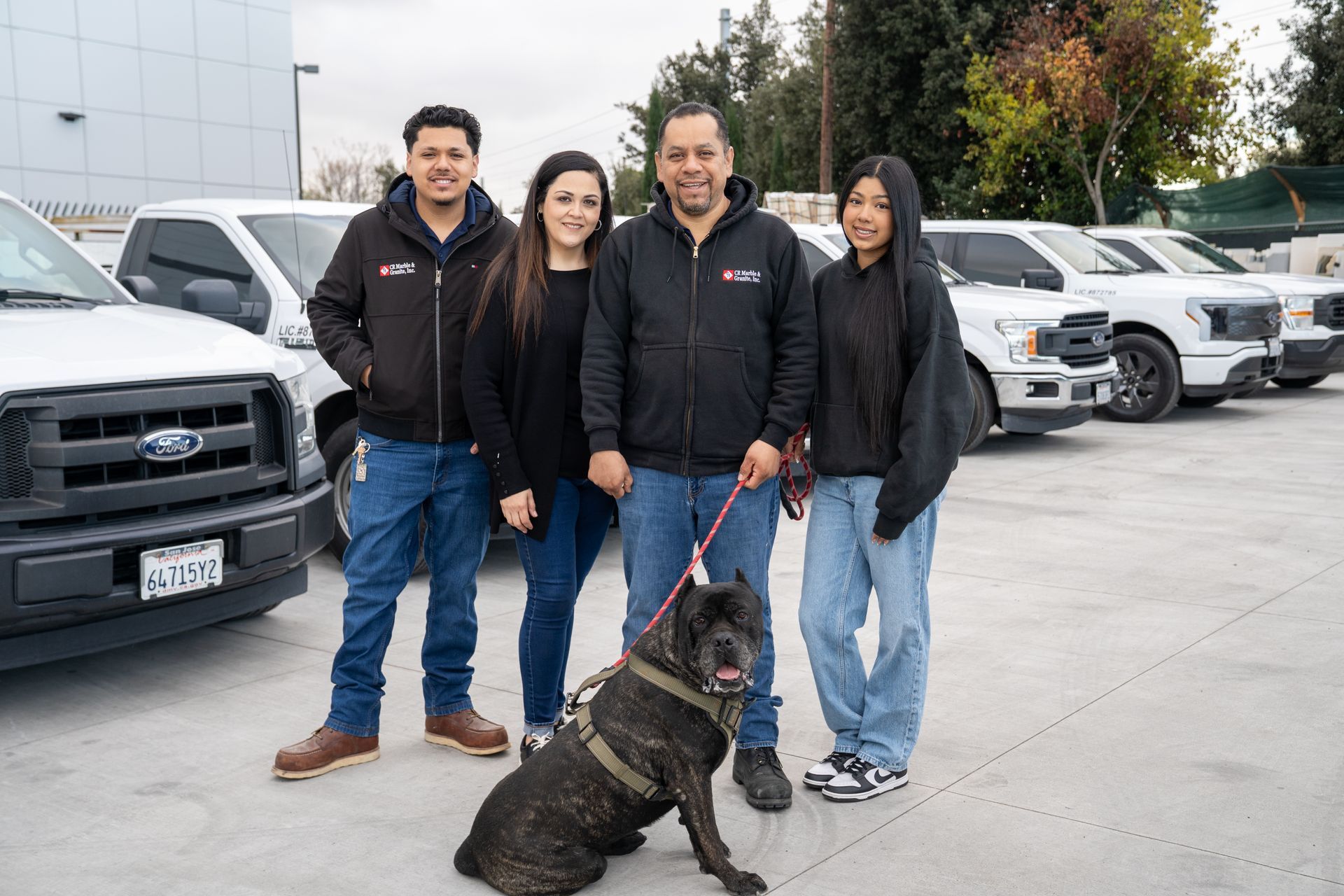 Four people with a dog standing in a car dealership lot beside parked trucks