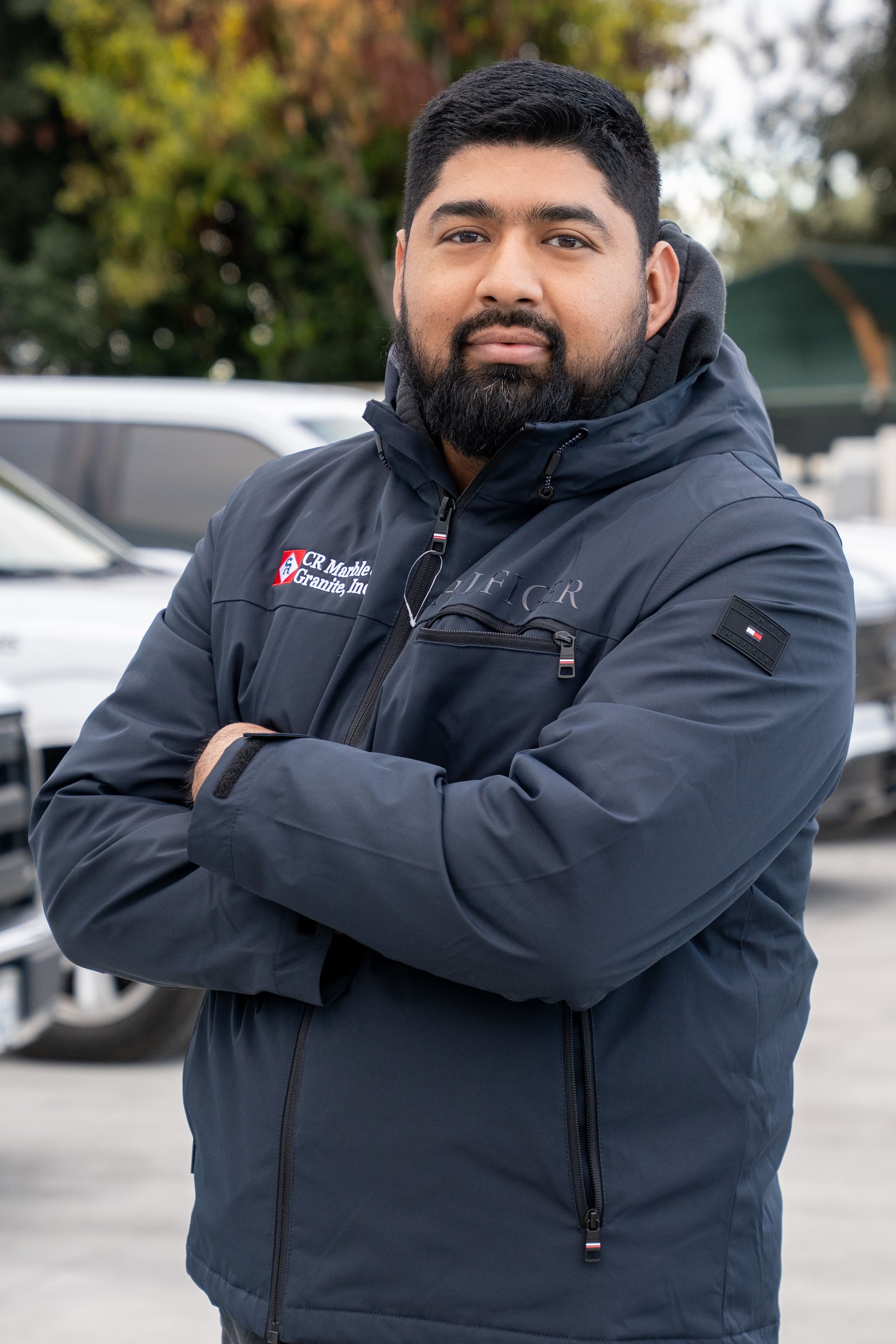Man in a dark jacket standing outdoors with arms crossed, in front of parked cars.