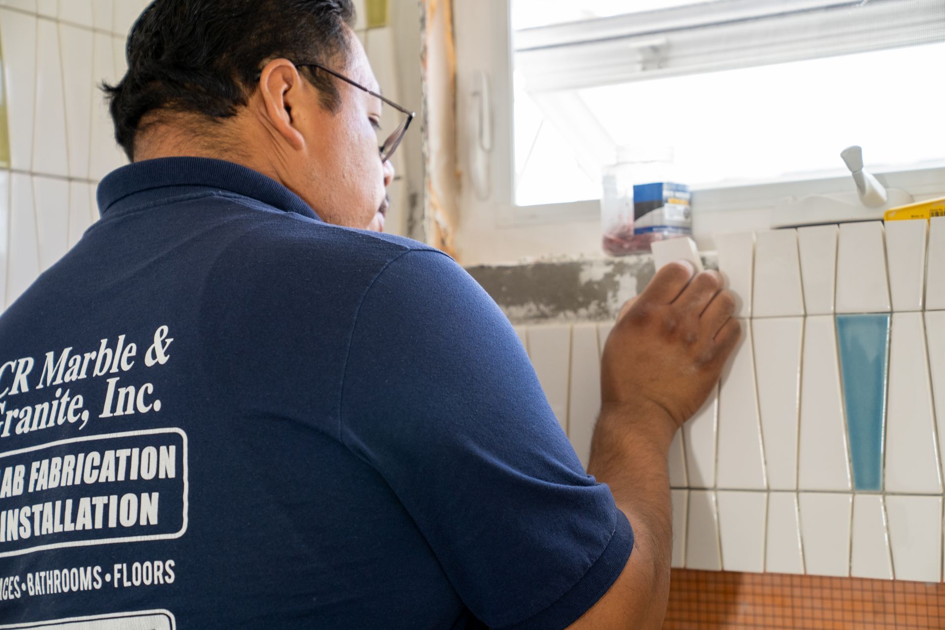 Worker installing white wall tiles in a bathroom or kitchen, holding a blue tile panel.