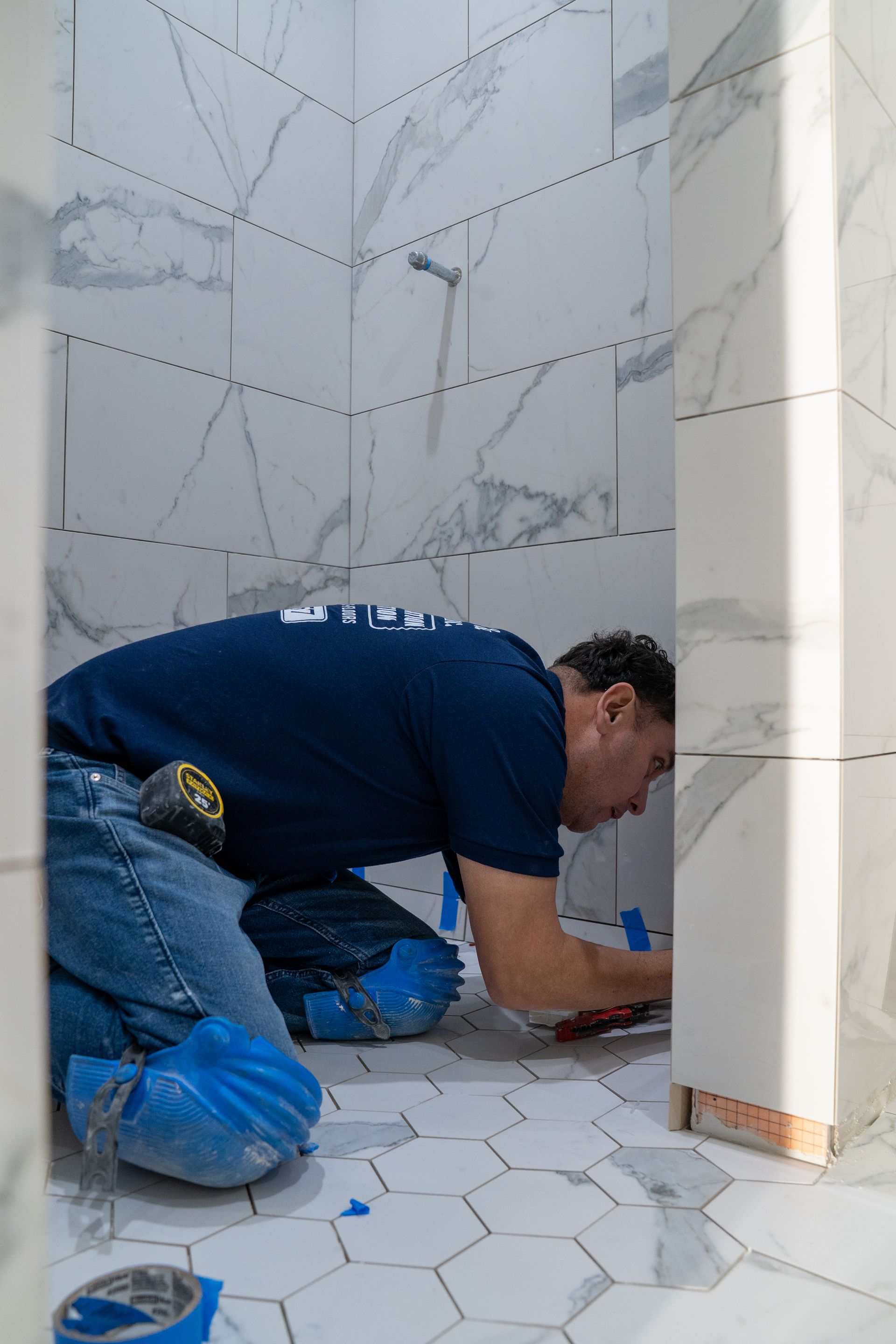 Worker installing tile in a white bathroom, kneeling near a corner wall.