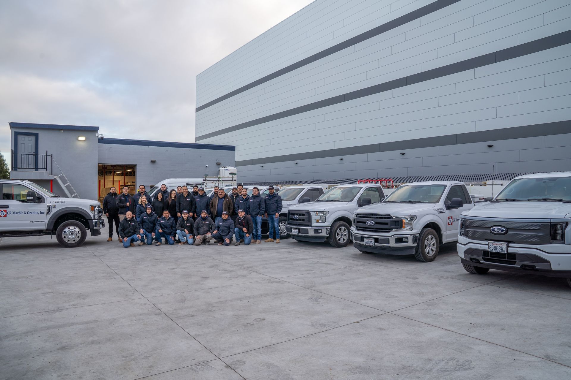 Group photo of people and trucks outside a modern industrial building at dusk