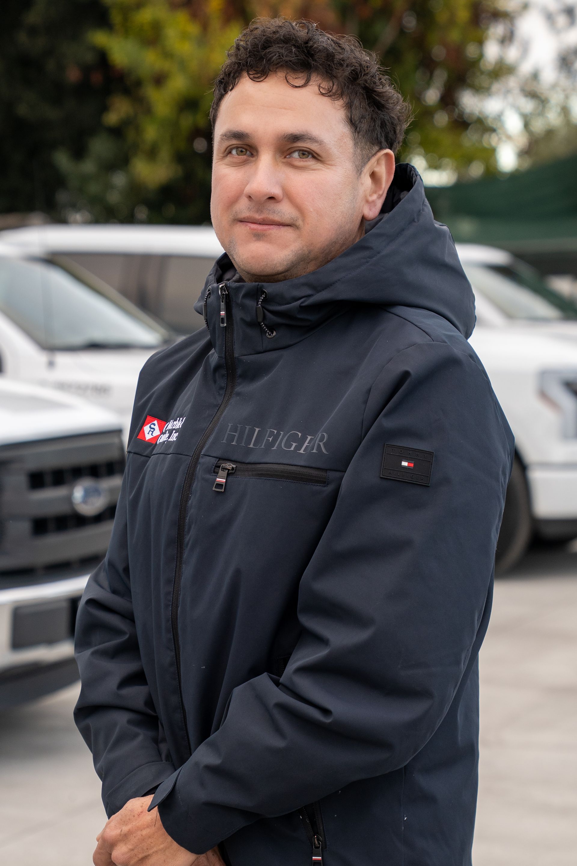Person in a dark jacket standing outdoors in front of parked cars, looking at the camera.