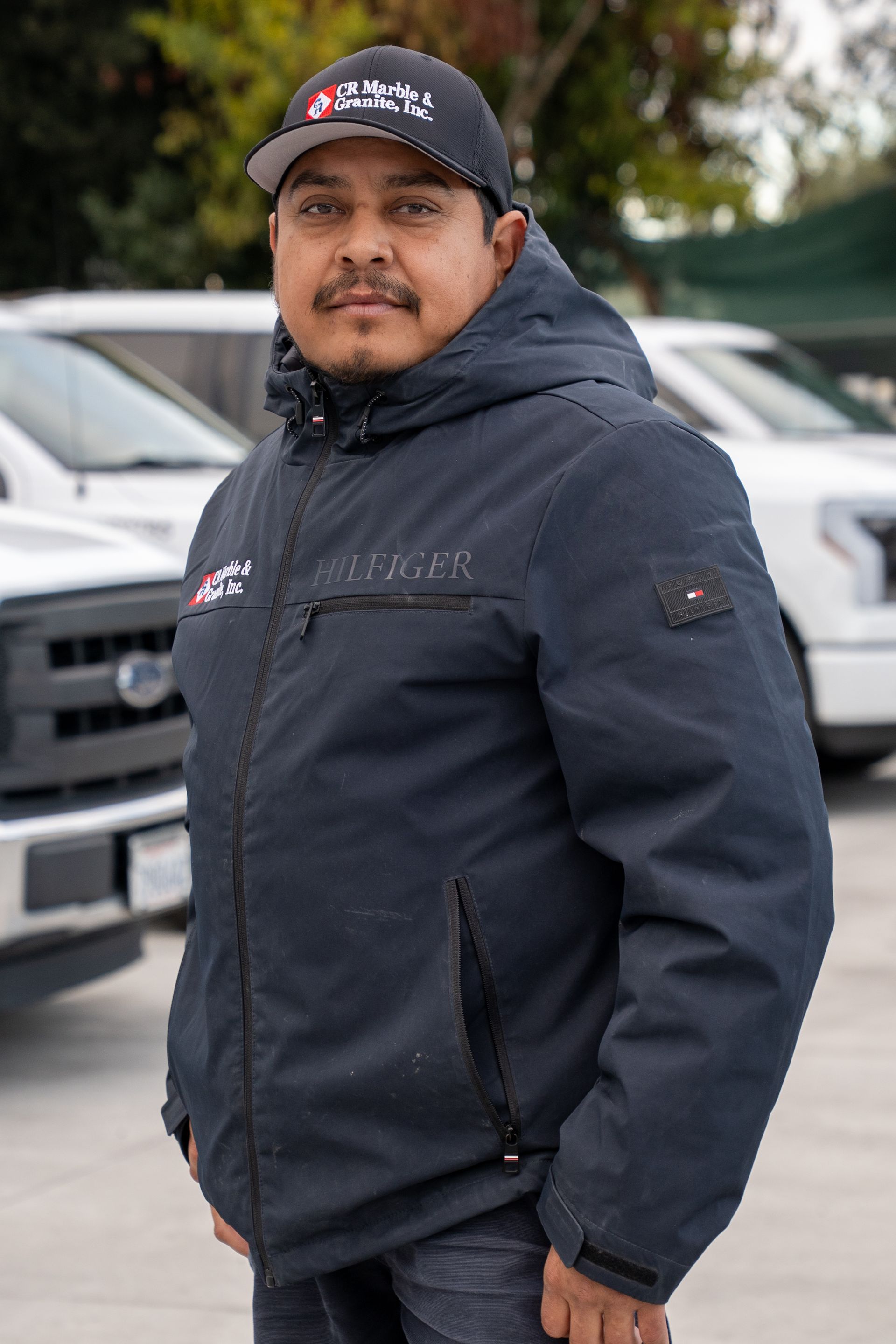 Man in dark jacket and cap standing outdoors near parked cars.
