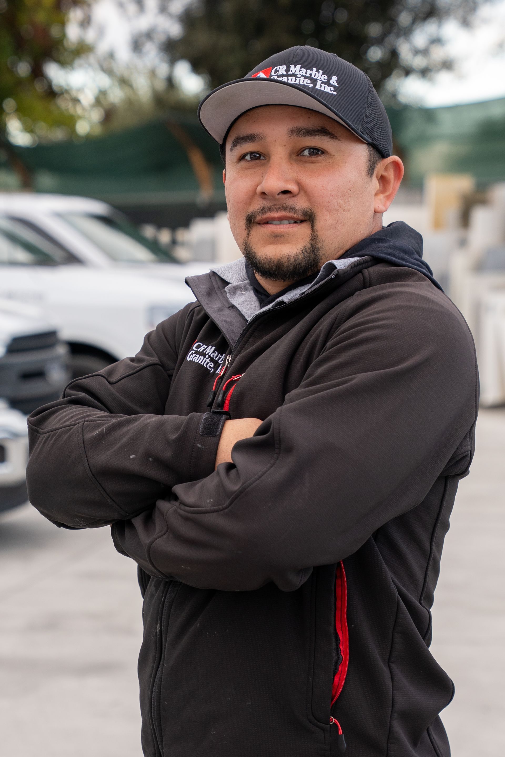 Person in a black jacket and cap standing with arms crossed outdoors, with cars blurred in the background