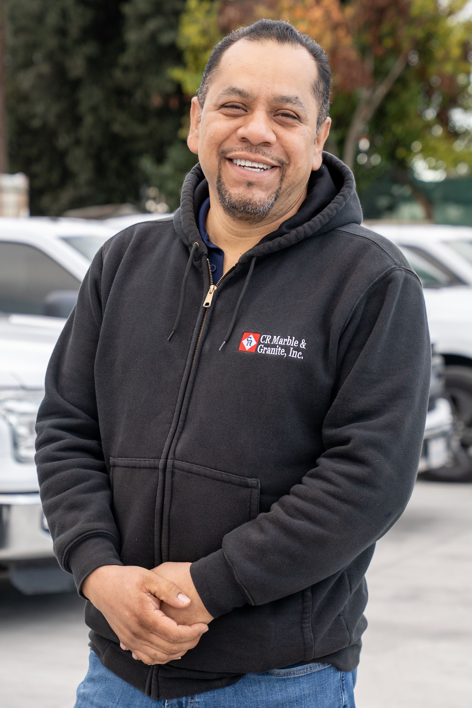 Smiling man in a black hoodie standing outdoors with parked cars in the background