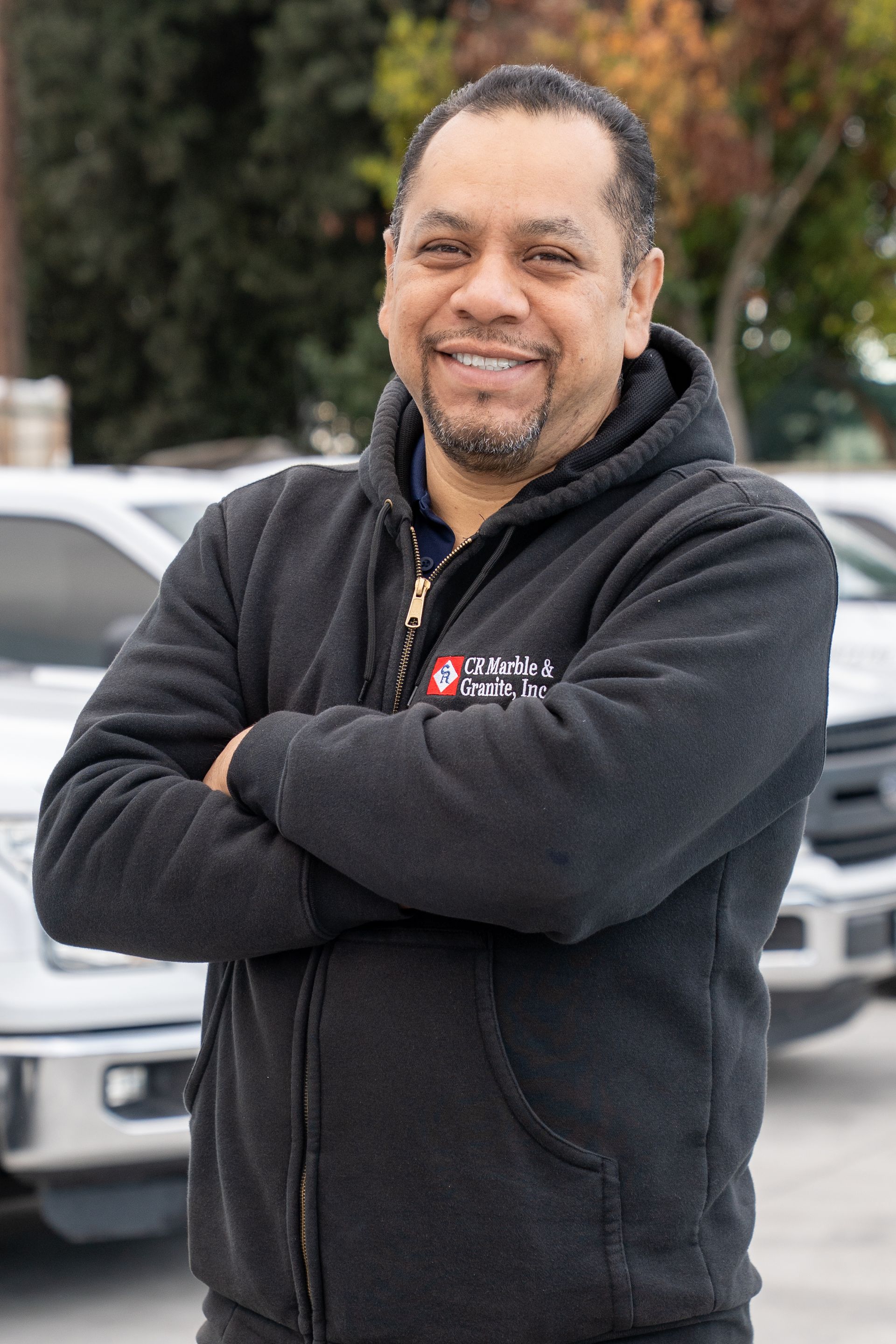 Smiling person in a black jacket standing with arms crossed outdoors in front of cars