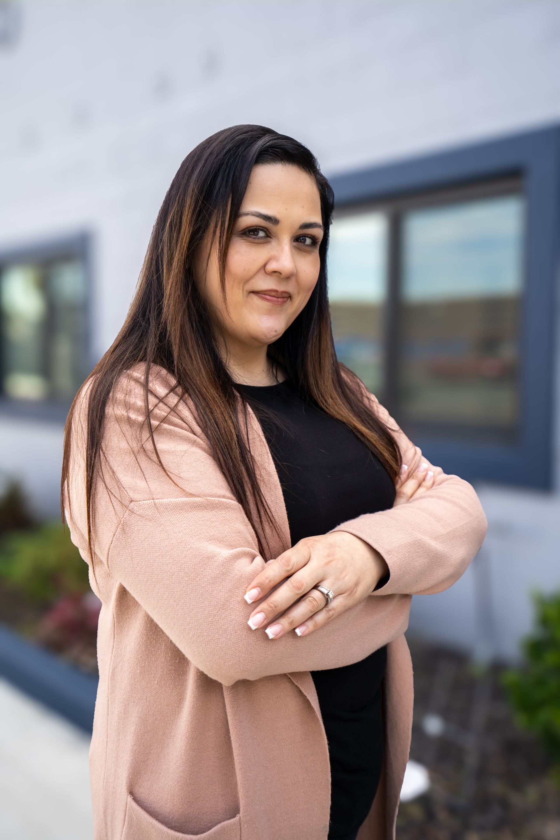 Woman with long dark hair standing outdoors with arms crossed in a pink cardigan and black top