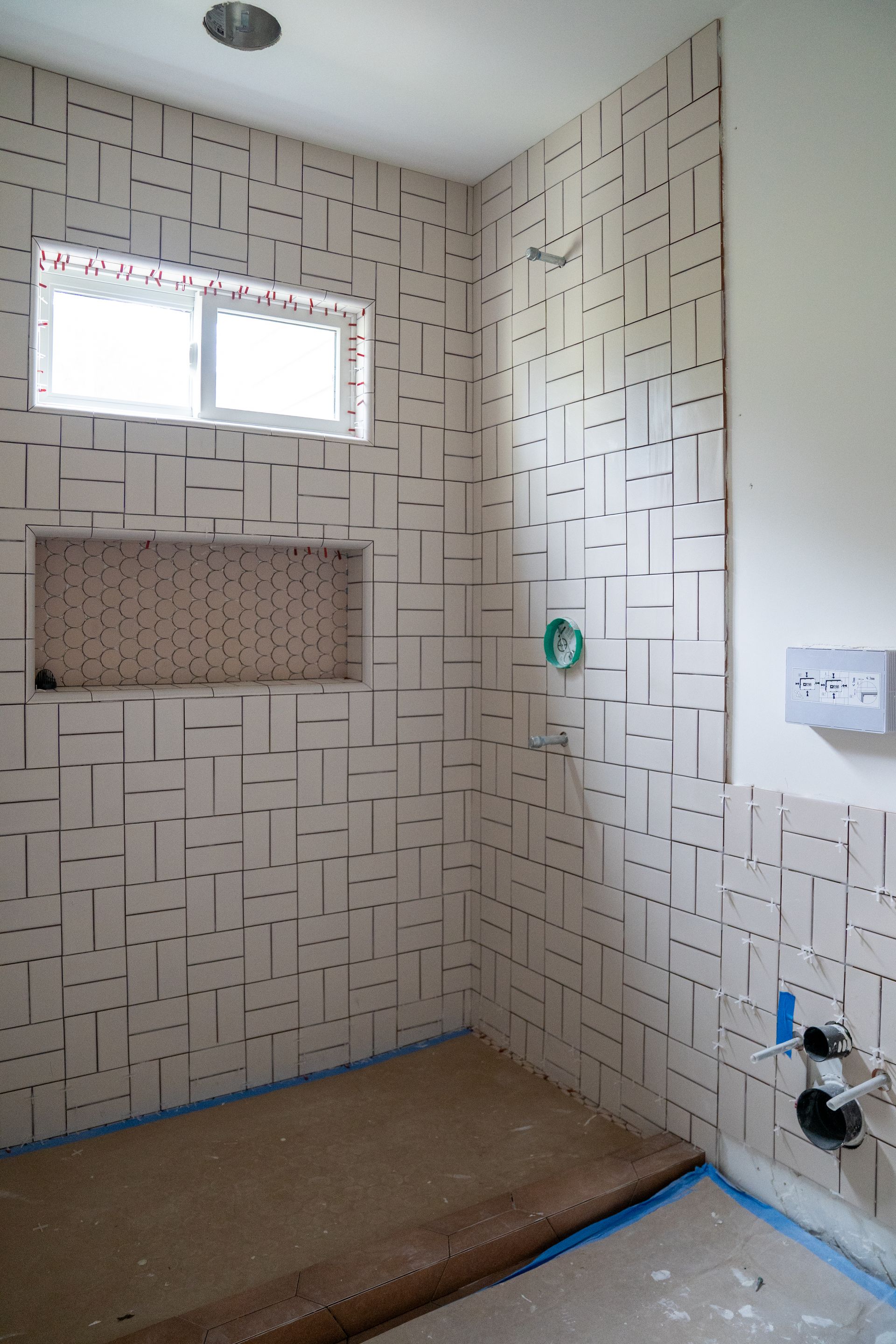 Tiled bathroom under renovation with a small window, shower plumbing, and blue tape around the floor edges