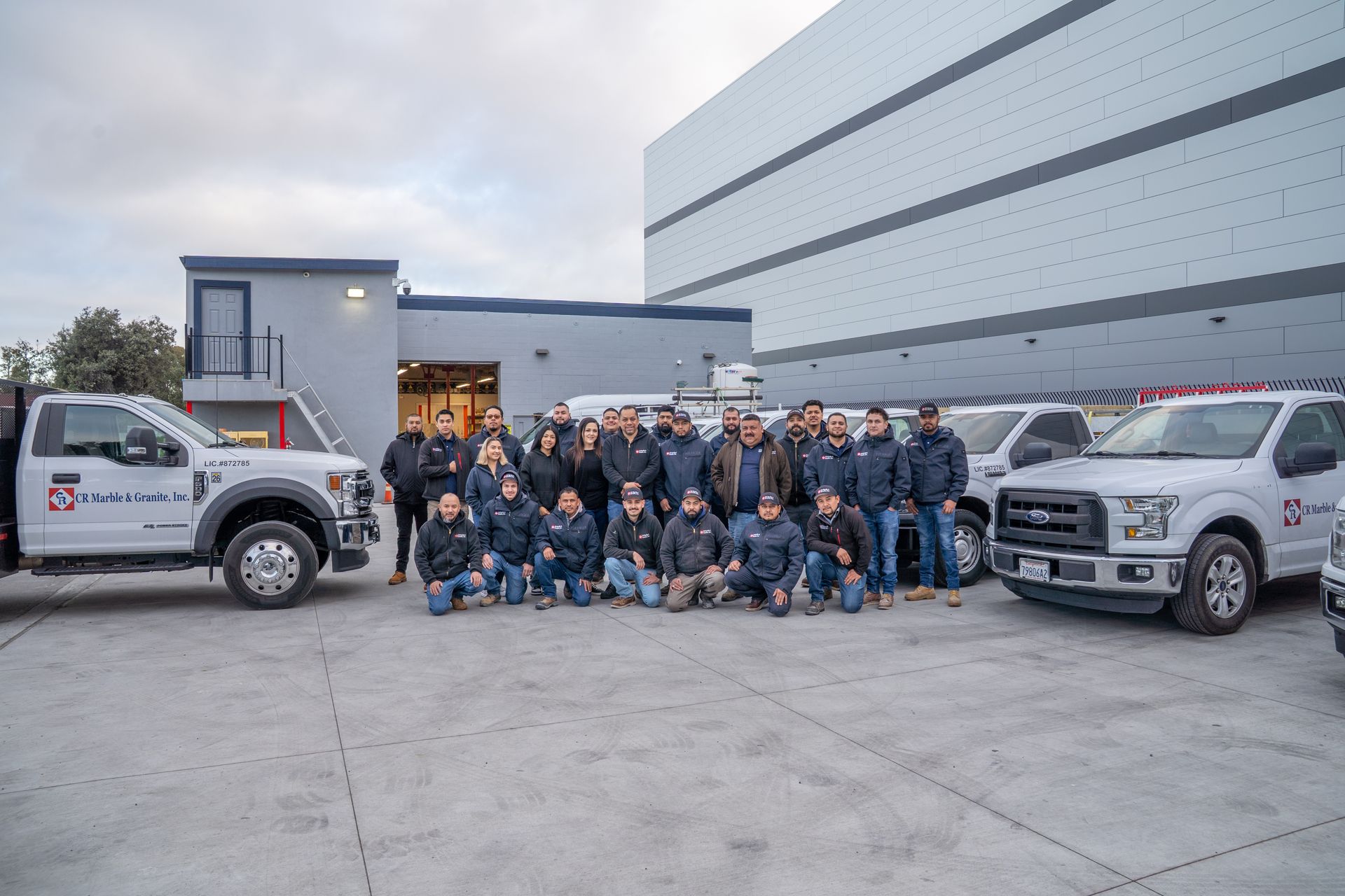 Large group posing outside an industrial building beside two white pickup trucks.