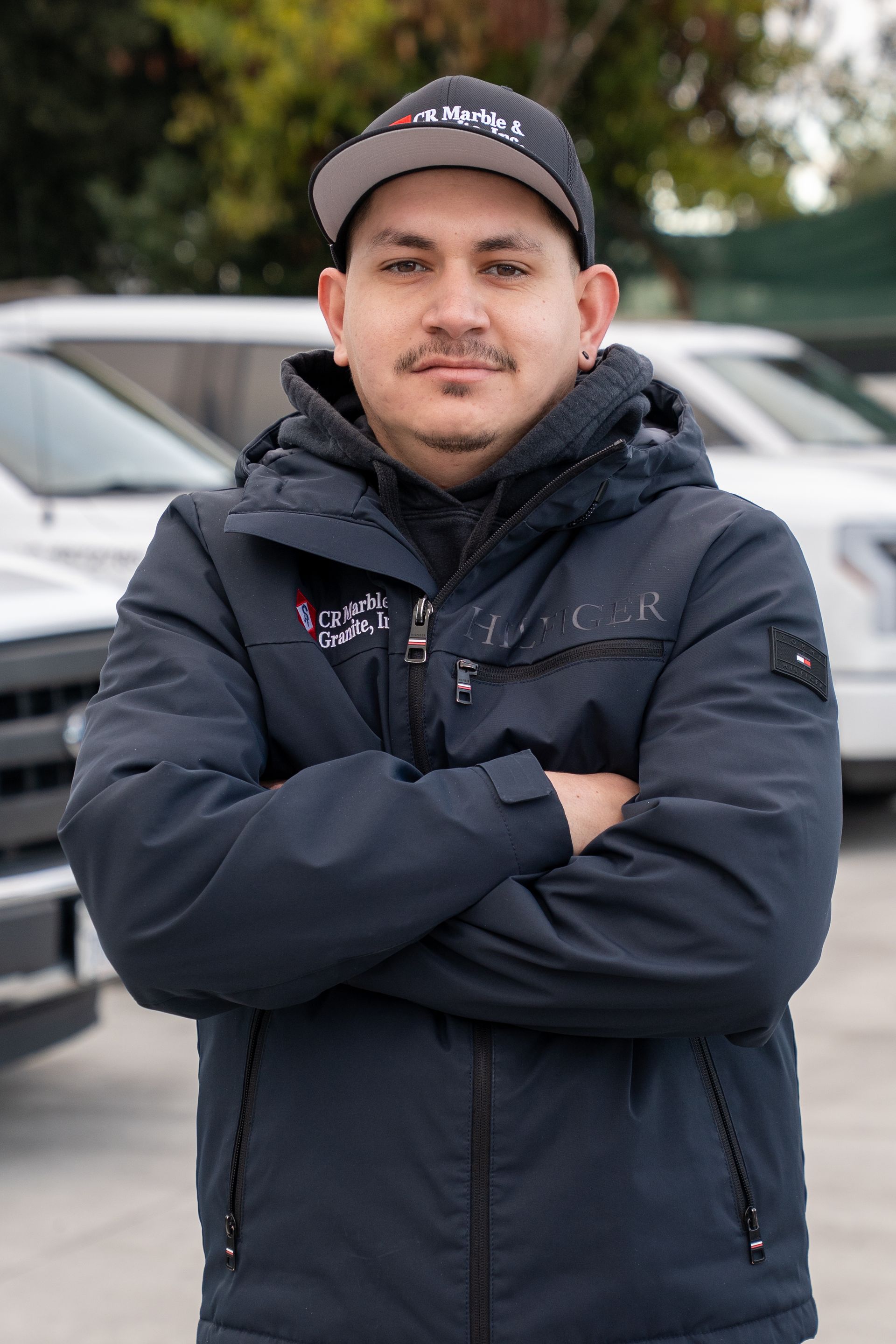 Person in a black jacket and cap stands with arms crossed in front of parked cars.