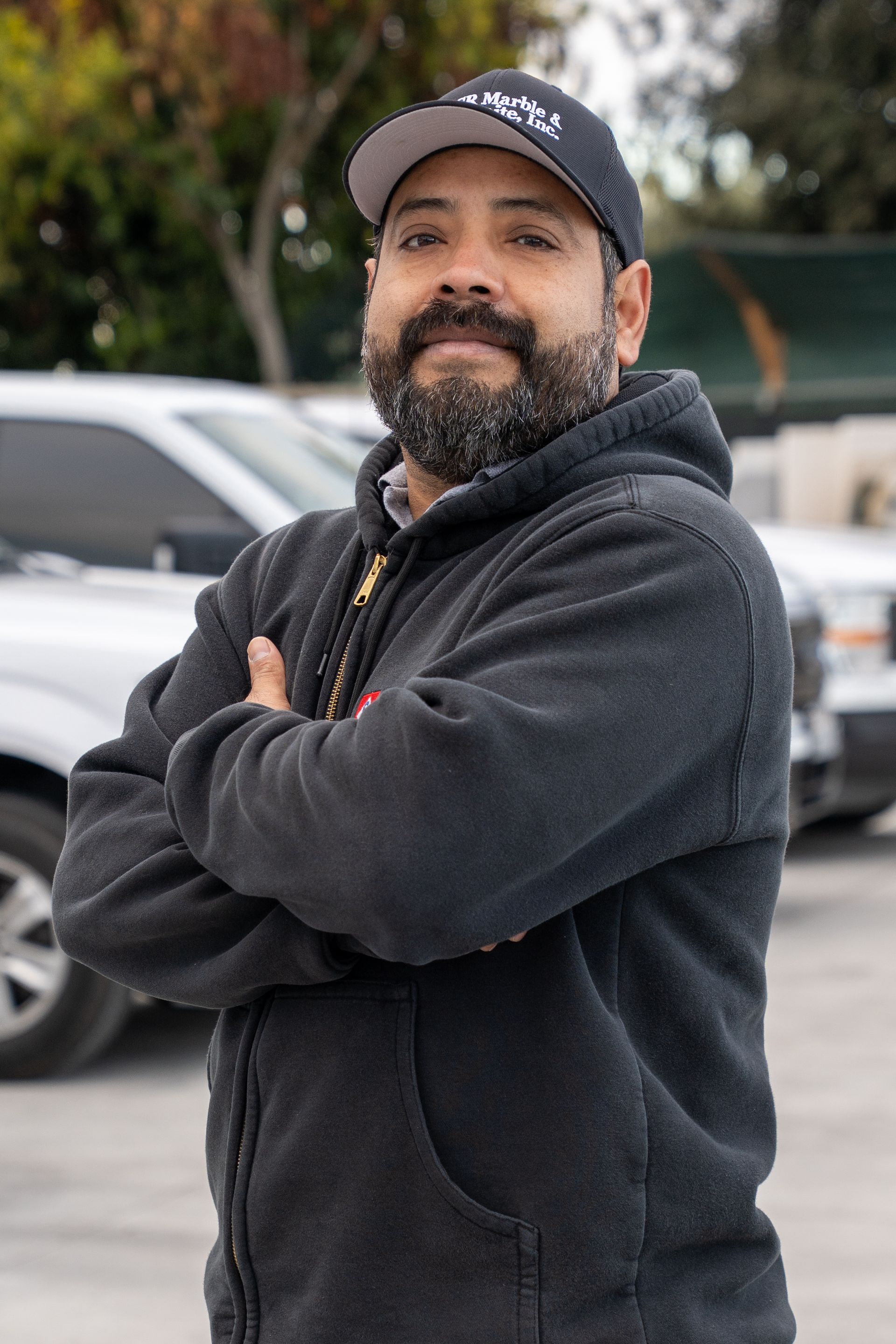 Smiling man in a black hoodie and cap standing with arms crossed outdoors near parked cars