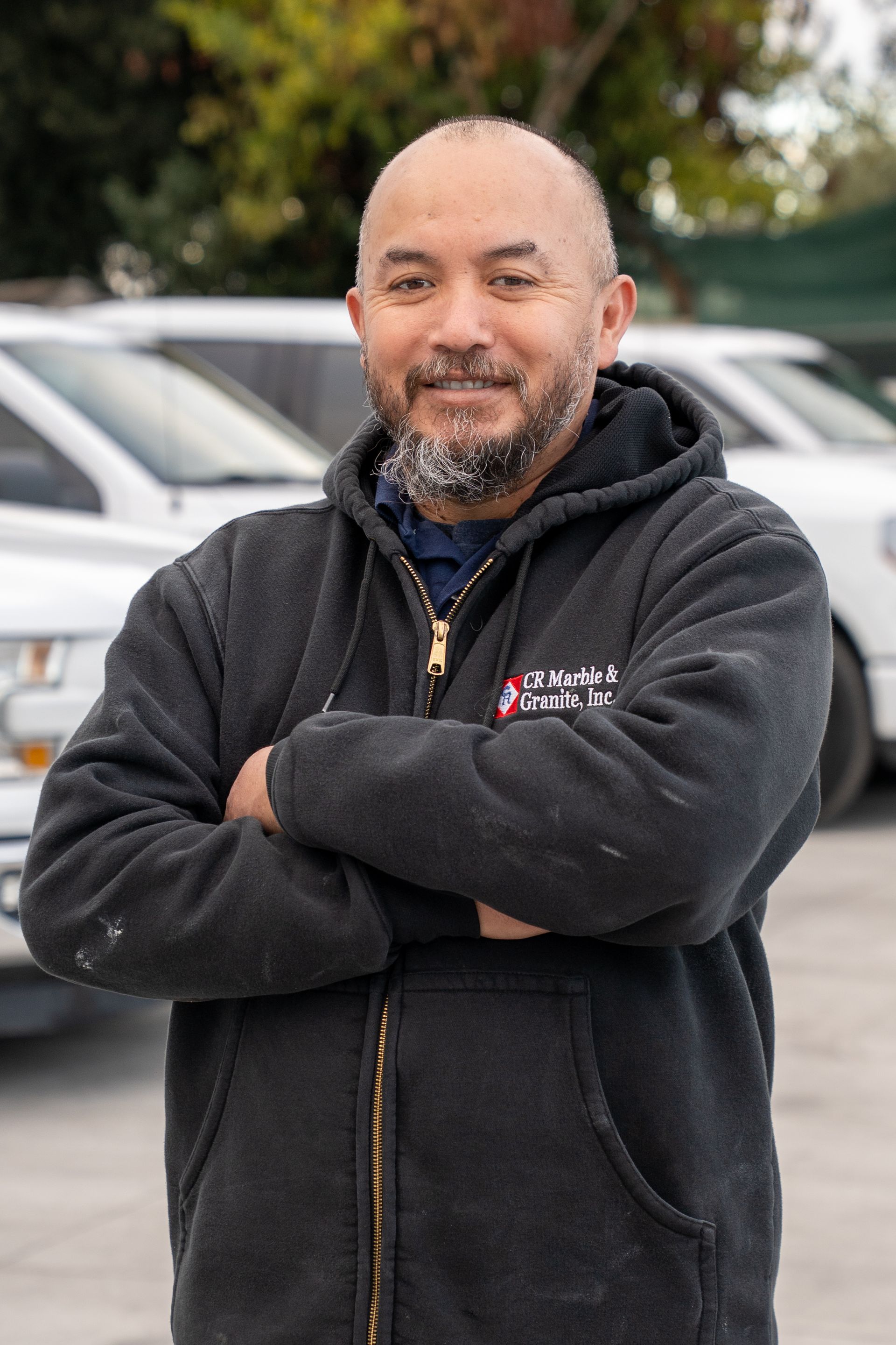 Man in a black hoodie standing with arms crossed in front of parked cars, smiling slightly.
