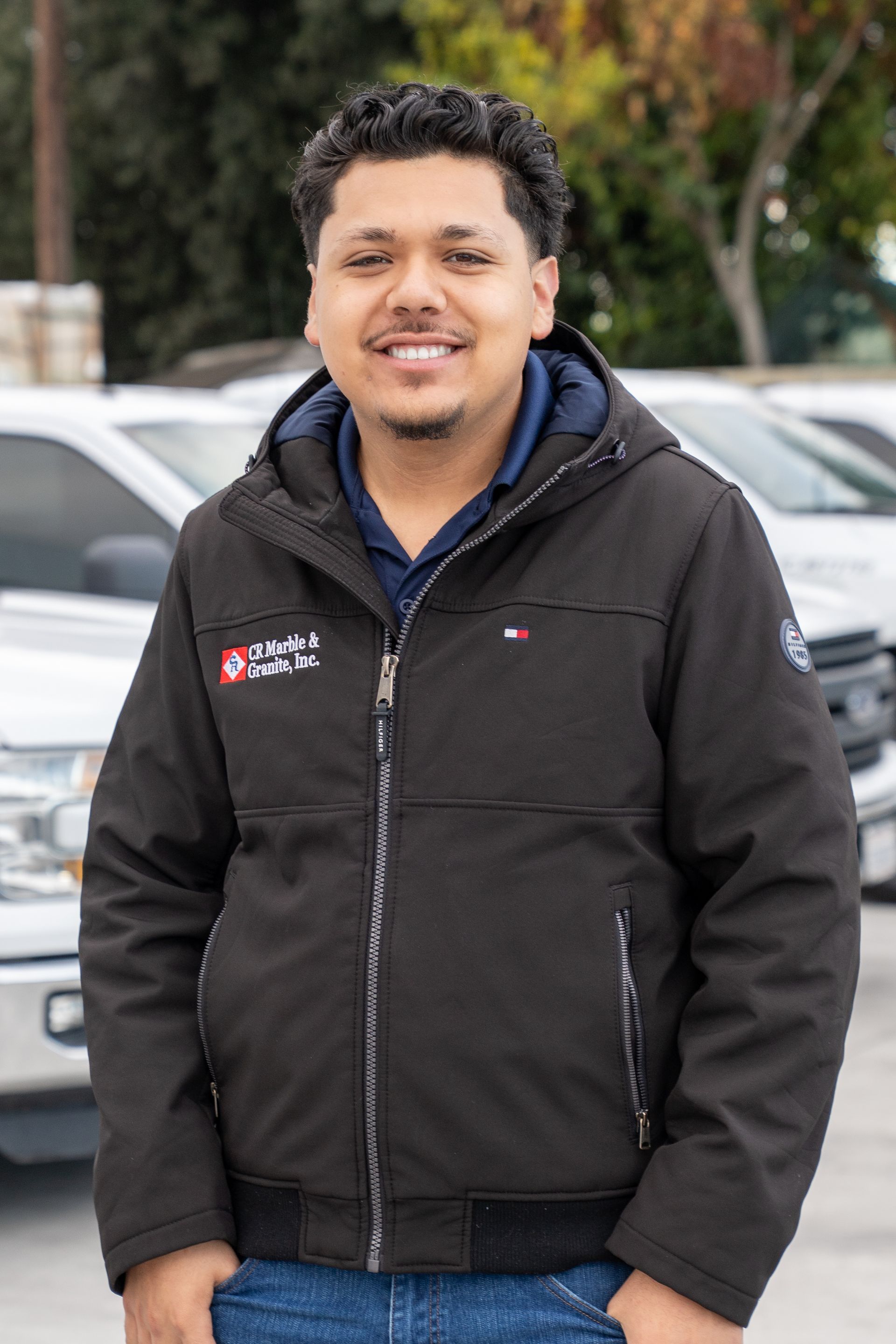 Man smiling in a black jacket standing outdoors in front of parked cars