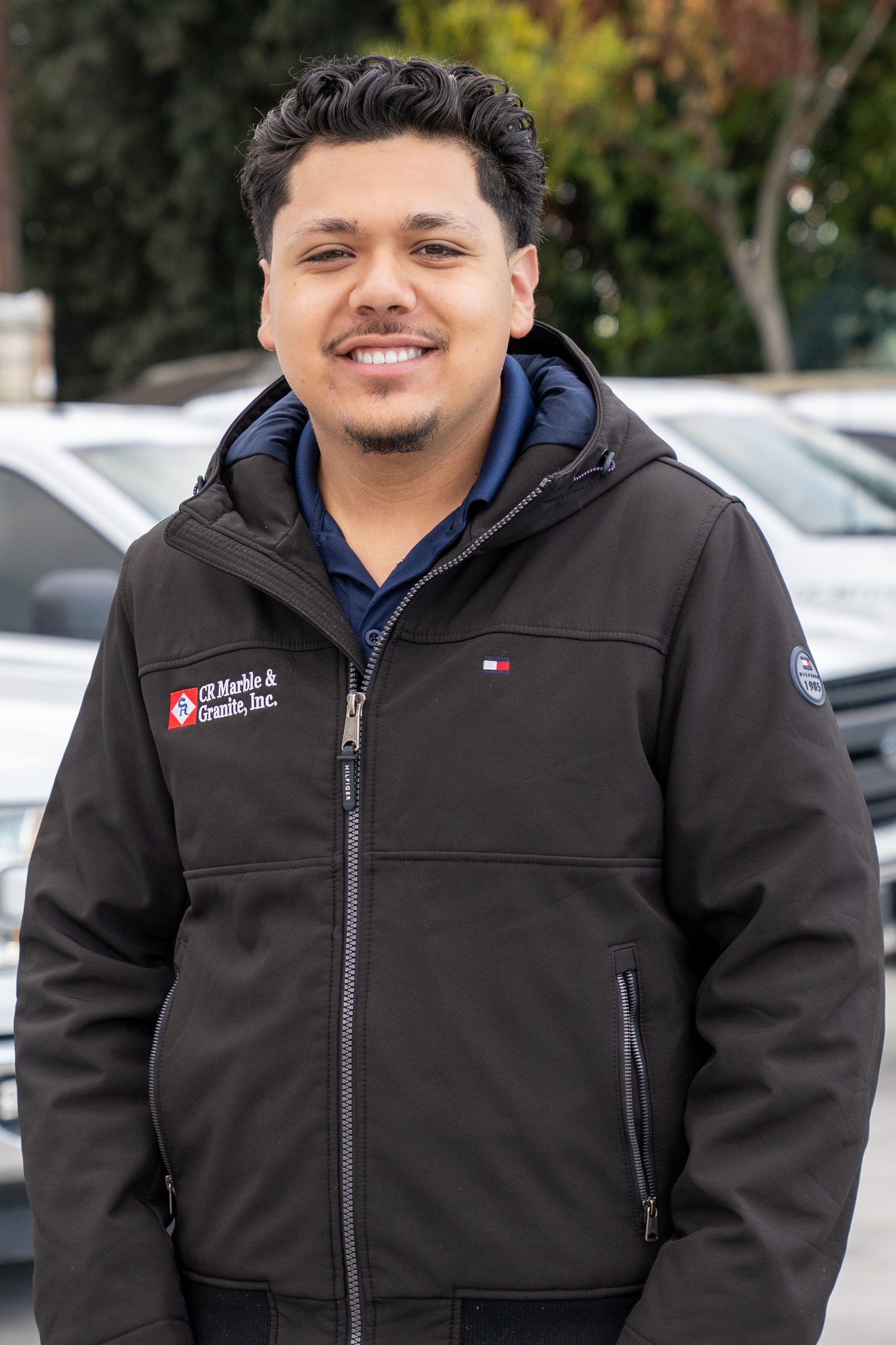 Smiling person in a black jacket standing outdoors in front of parked cars and trees