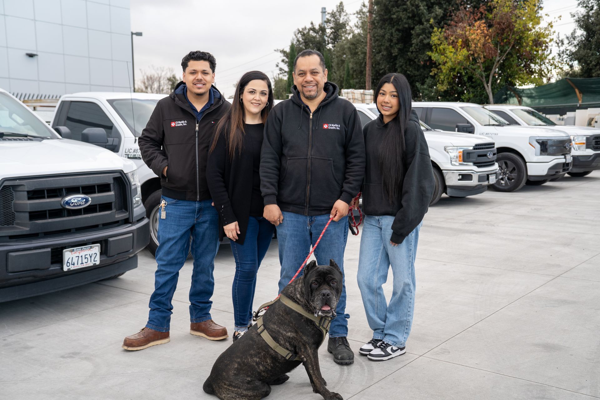 Four people and a dog standing in a car dealership lot with parked trucks behind them