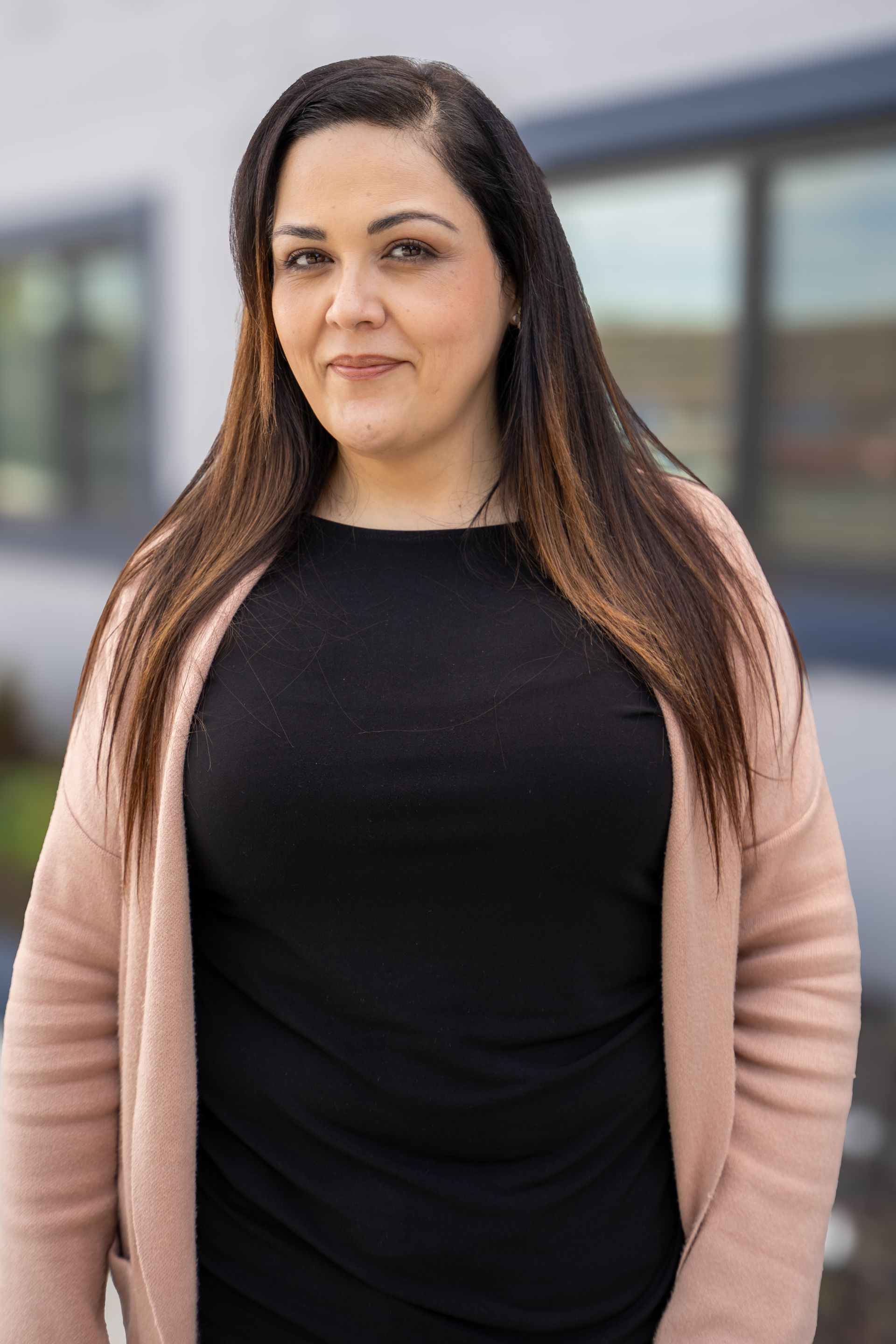 Woman standing outdoors in a black top and pink cardigan, with a blurred building in the background