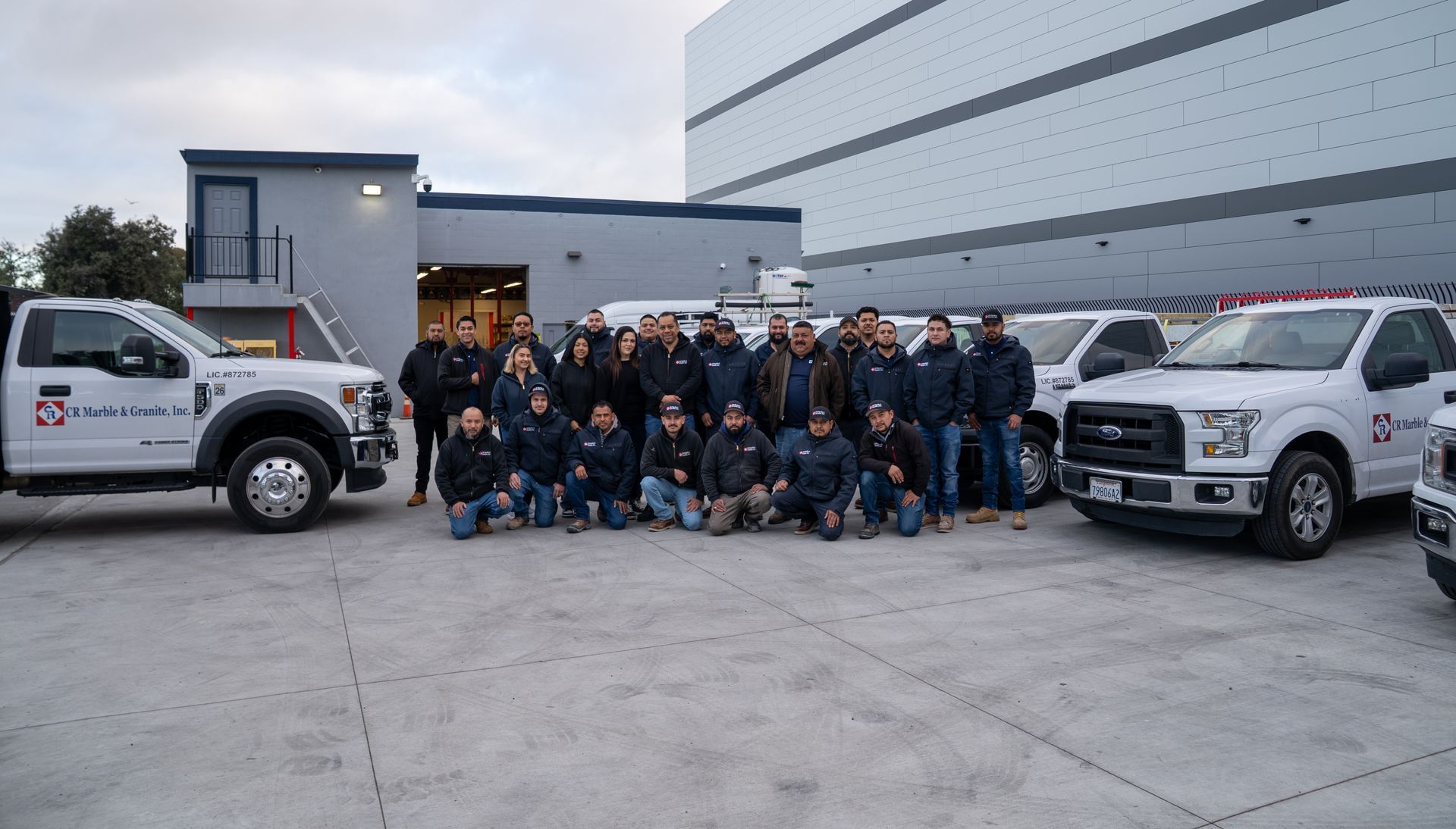 Group of workers posing in front of two white utility trucks beside a large industrial building