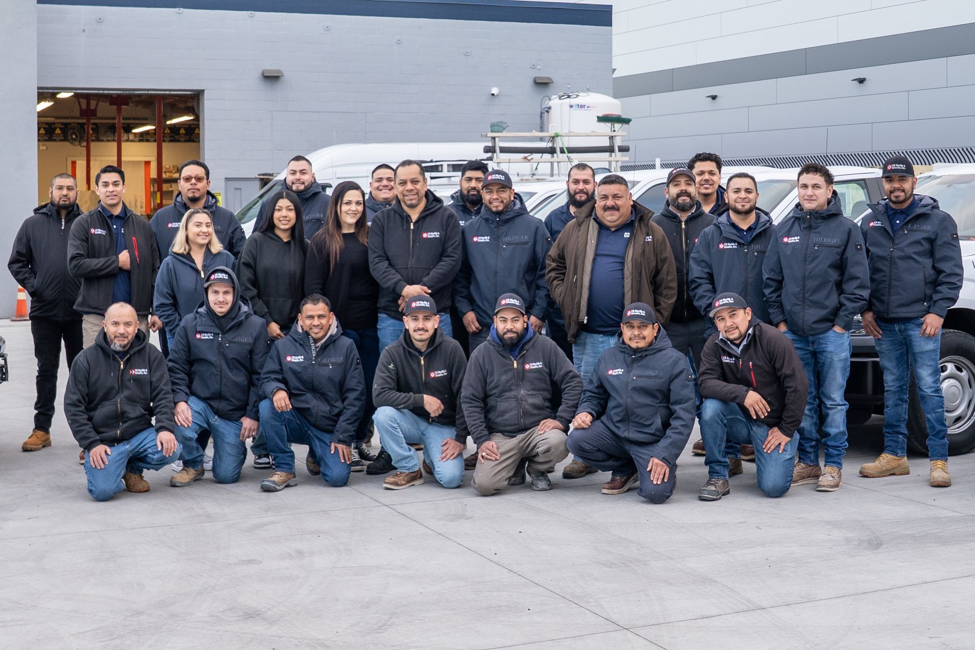 Group of people in dark jackets posing outside a gray building, with some kneeling in front.