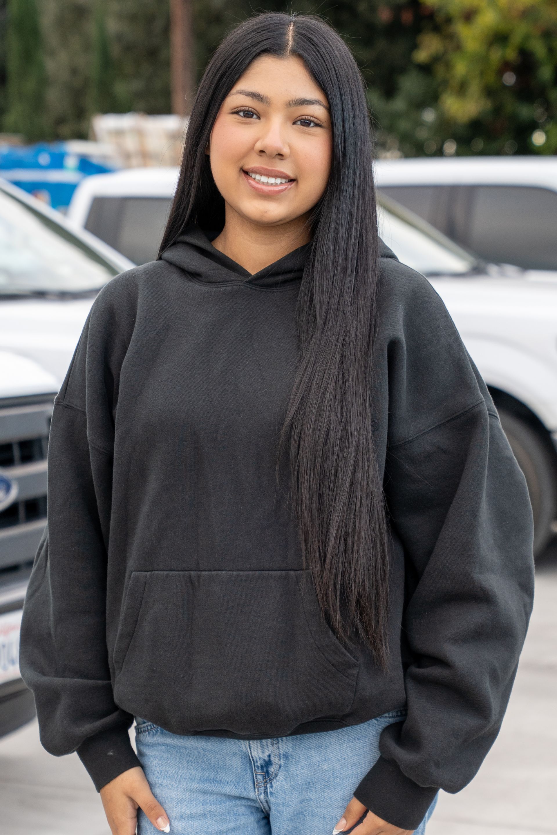 Person standing outdoors in a black hoodie and blue jeans, smiling slightly, with parked cars behind them