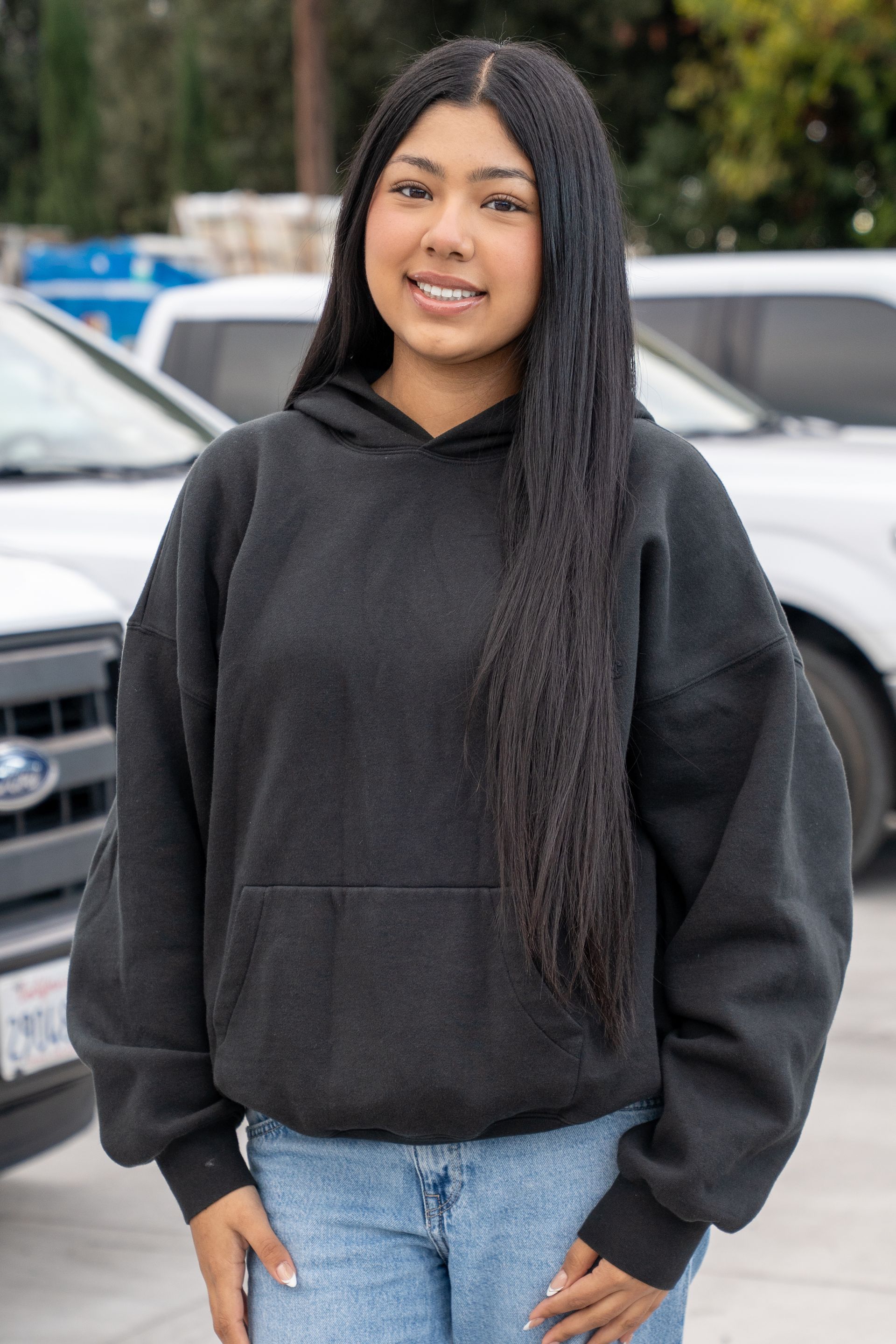 Person standing outdoors in a black hoodie and jeans, with parked cars in the background.